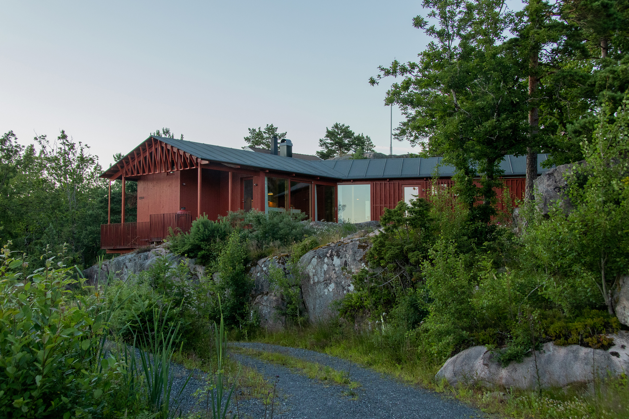 Red cabin with a trussed porch roof perched above rock outcrops and pines, reached by a gravel path in summer