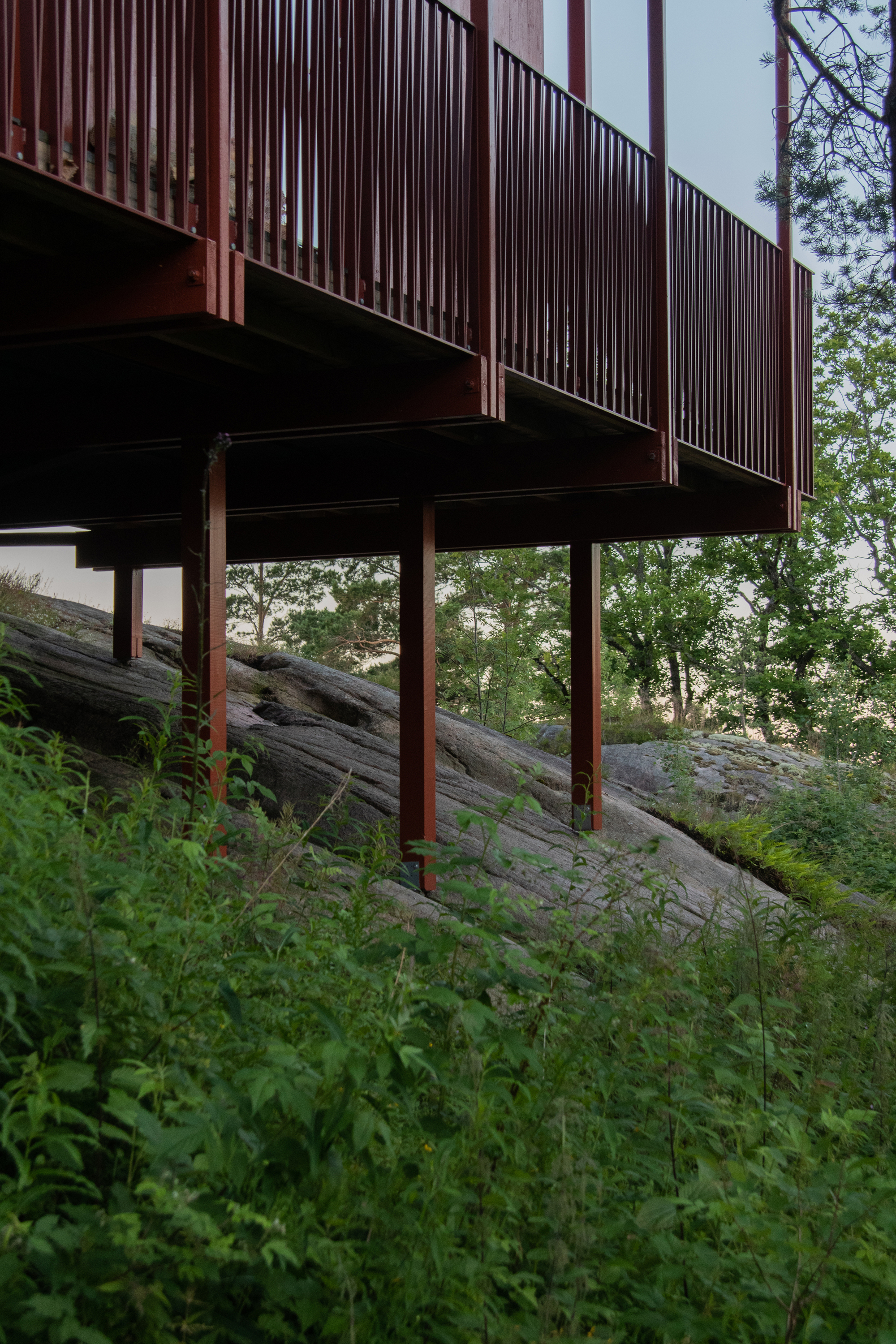 Underside of the red steel-framed deck raised on slender posts above a sloping granite outcrop, with green undergrowth in the foreground