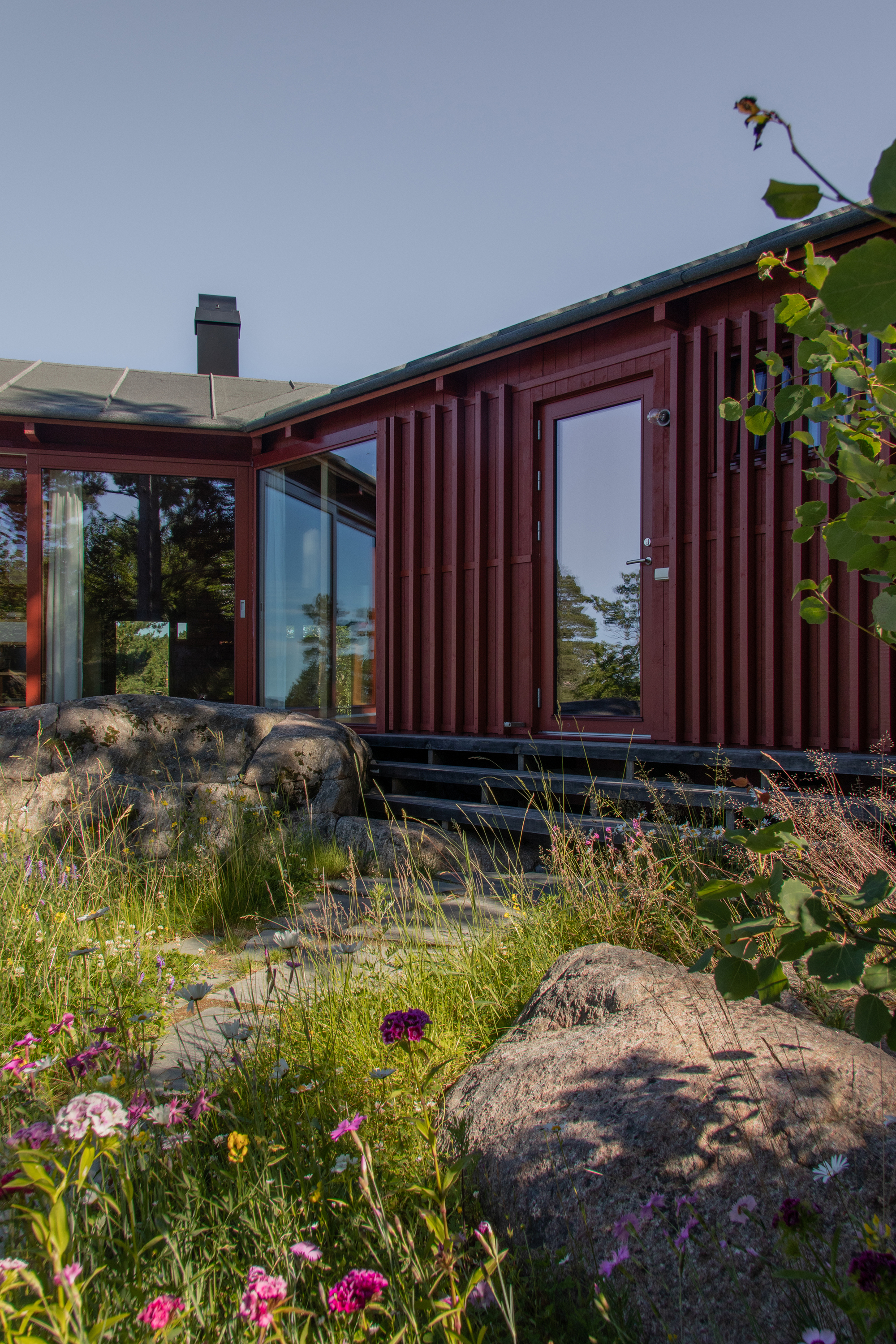 Red vertical-board cabin wall with glazed door and corner windows, set among boulders and wildflowers in summer light