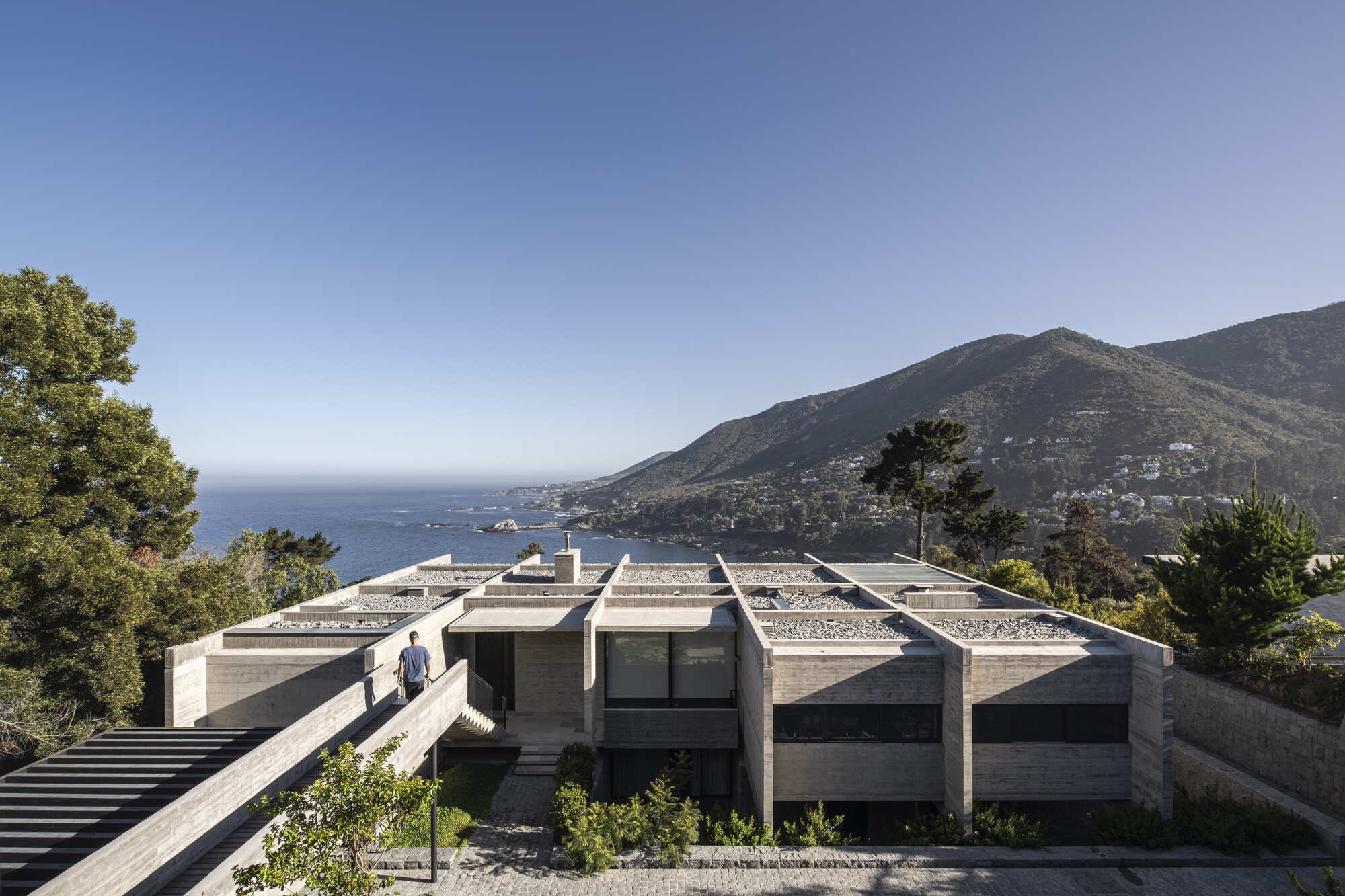 Aerial view of the board-formed concrete building with gravel roof bays, a man on the stair, and the ocean and mountain beyond in clear afternoon sky