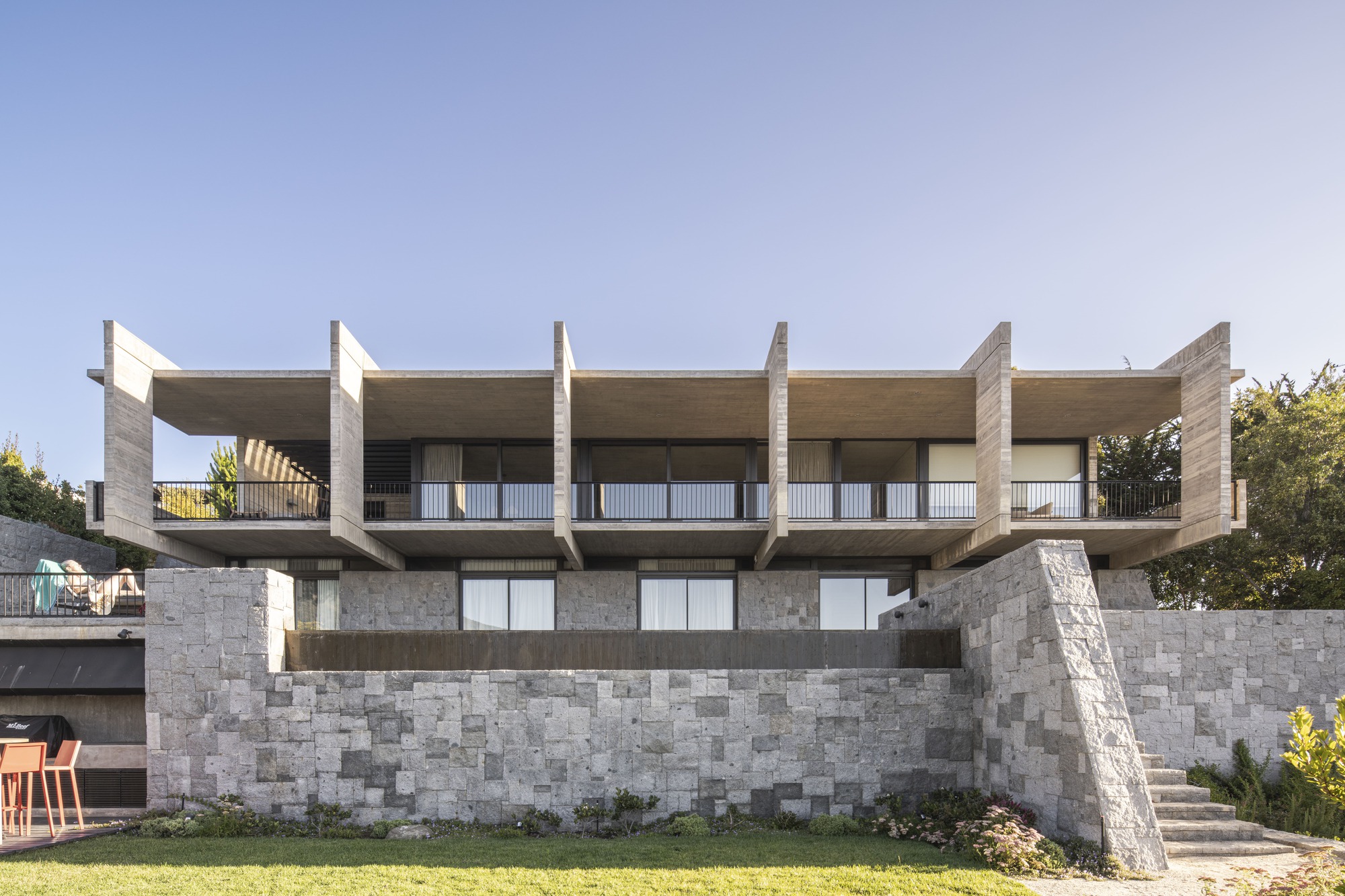 Garden facade with projecting concrete fins over a grey stone retaining wall and a lawn in late afternoon light