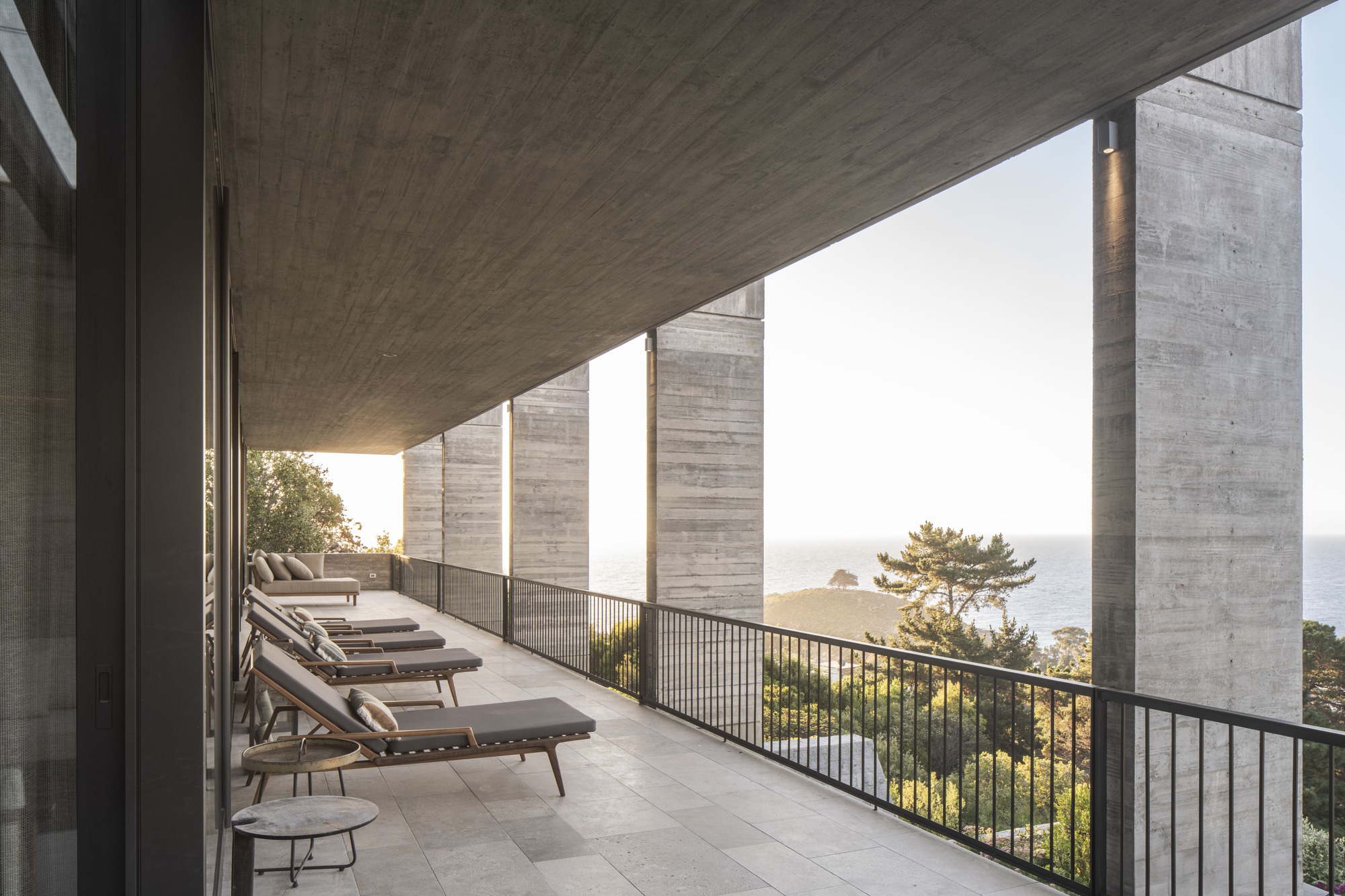 Covered terrace with lounge chairs along a black railing between concrete piers, the sea glowing at sunset