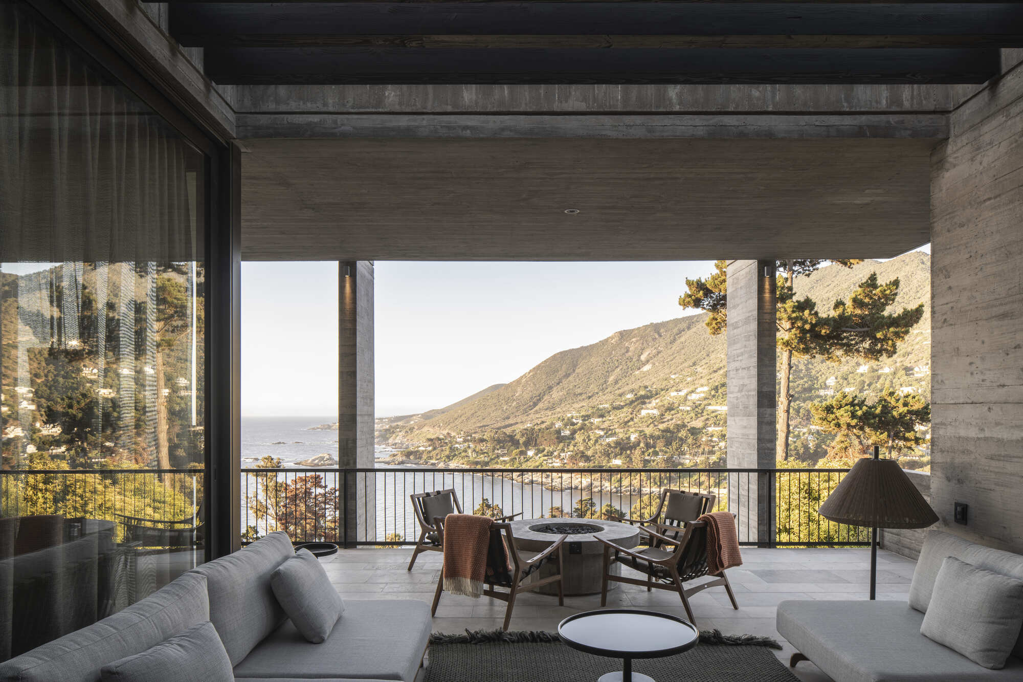 Covered outdoor sitting area with a fire pit, timber chairs and sofas framing a view of the coastal hills at dusk