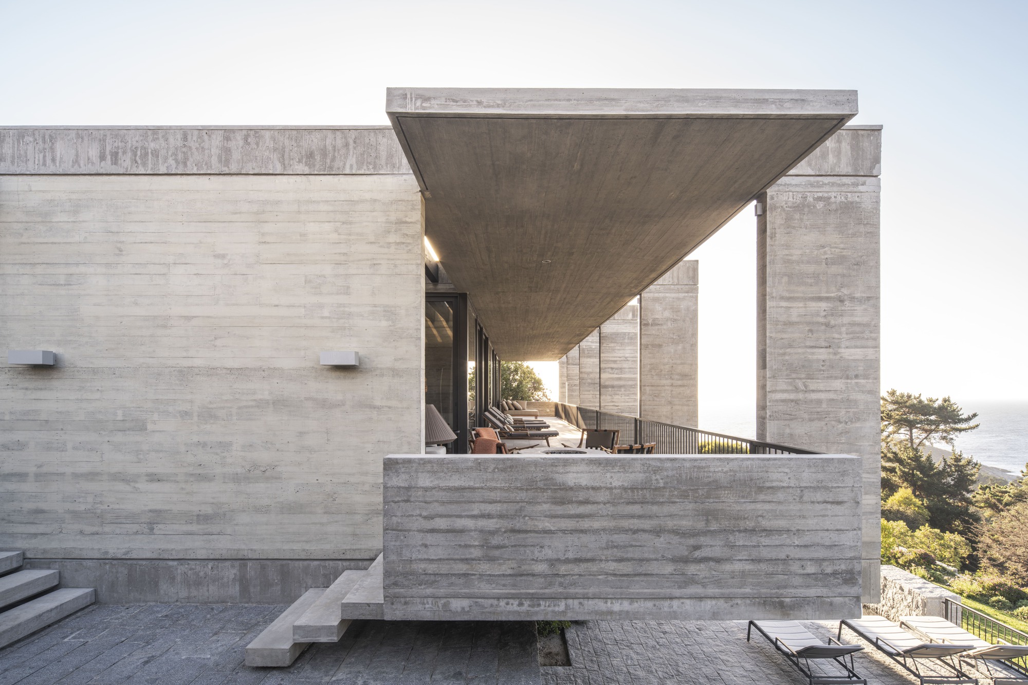 Side view of the concrete cantilevered canopy over the terrace, with stone steps and sun loungers in bright haze
