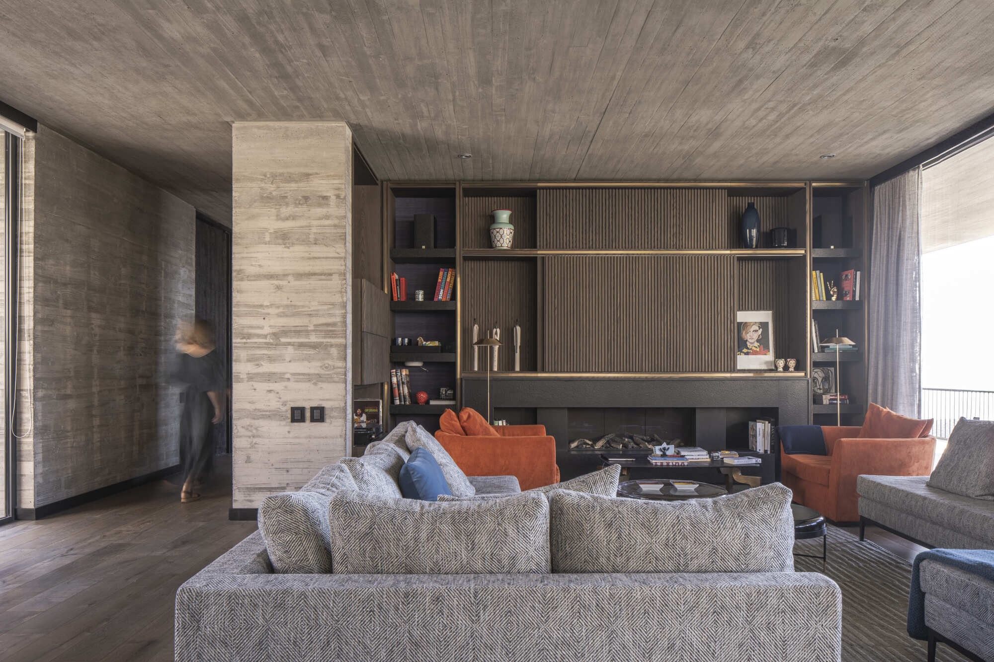 Living room with a grey sofa facing a slatted timber fireplace wall, a blurred figure walking past a concrete column