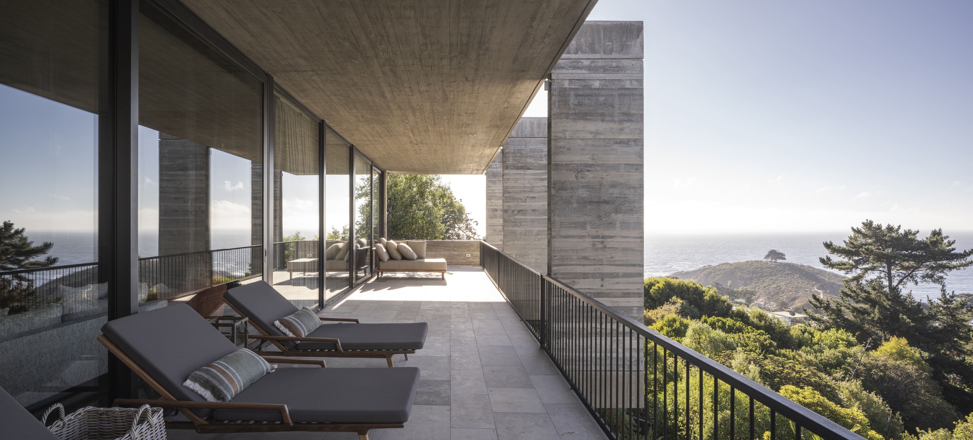 Terrace under a board-formed concrete soffit with grey lounge chairs facing a pine-covered headland and the ocean in clear afternoon light