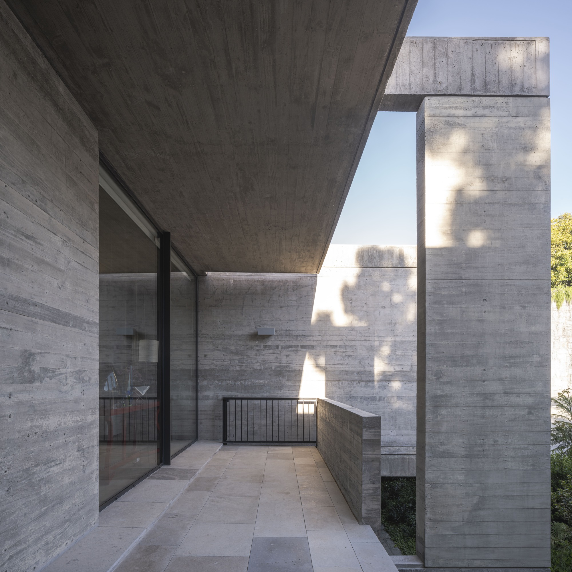 Corner of a concrete terrace with a freestanding column, steel railing and tree shadows falling across the board-marked walls