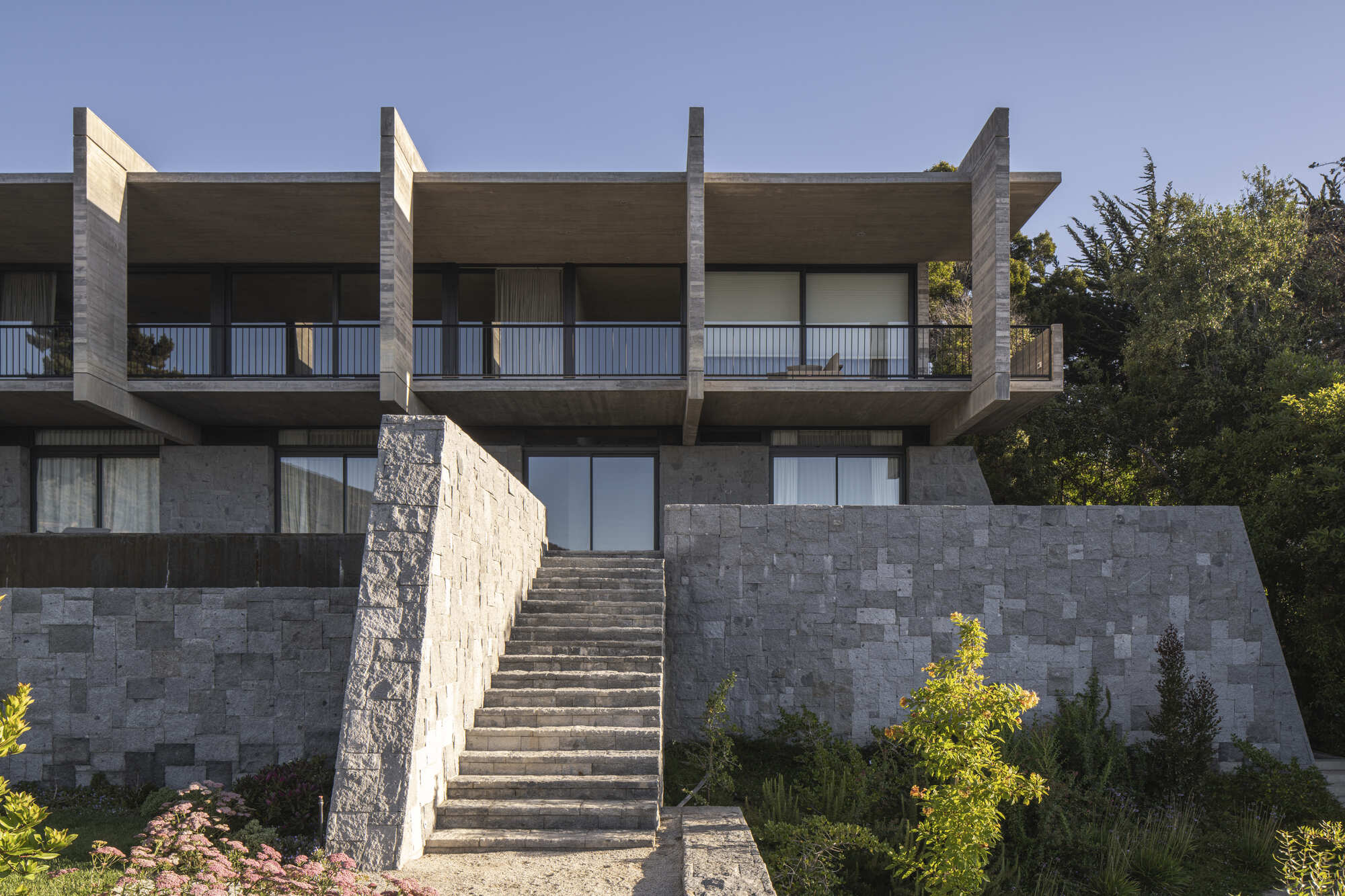 Stone stair rising between battered stone walls to a two-storey facade of concrete fins and balconies, with shrubs in the foreground