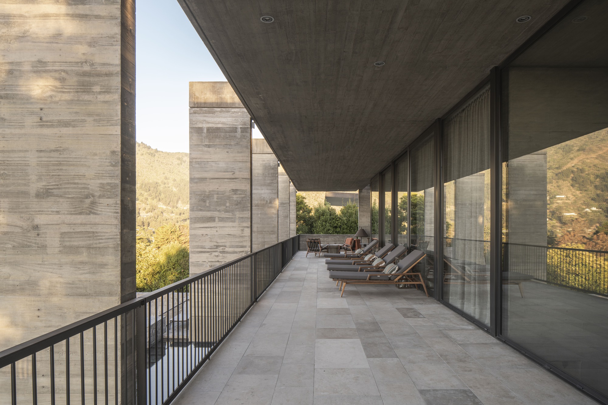 Long covered balcony with a row of lounge chairs beside floor-to-ceiling glazing and concrete piers, hillside trees beyond