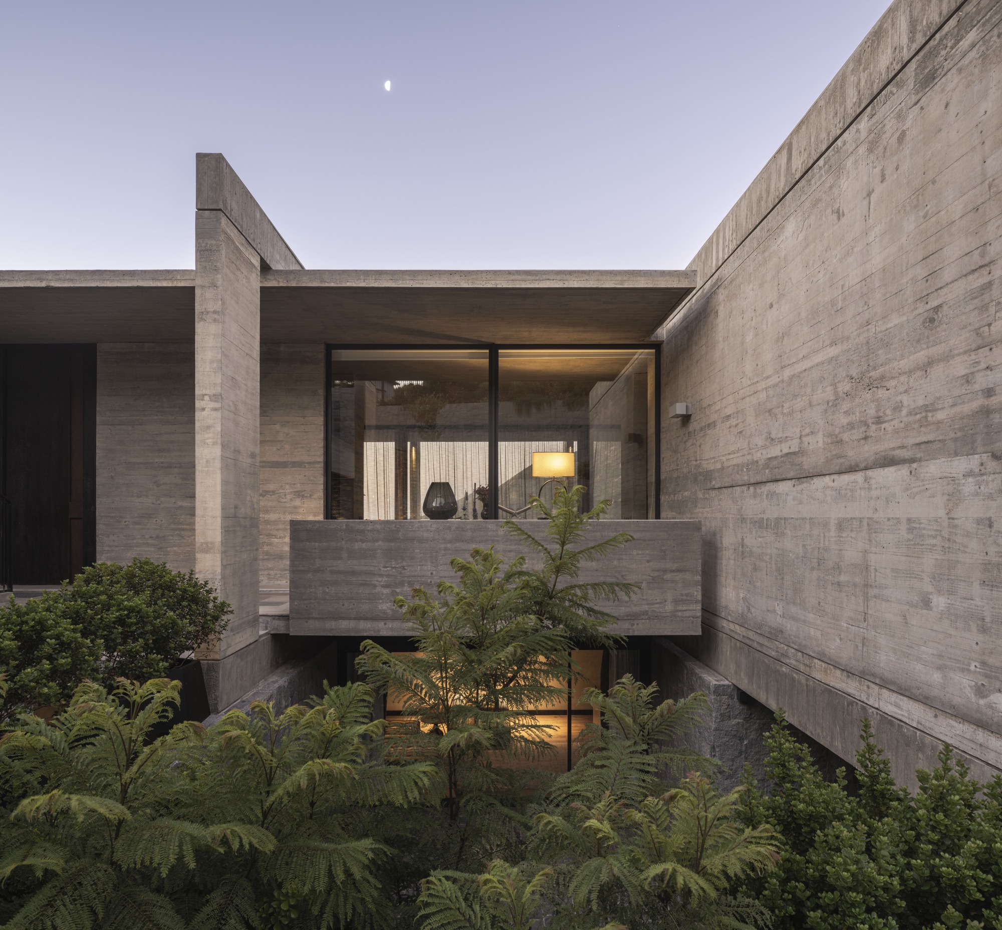 Cantilevered concrete balcony above a fern-filled courtyard at dusk, a lit lamp glowing in the window and a crescent moon overhead