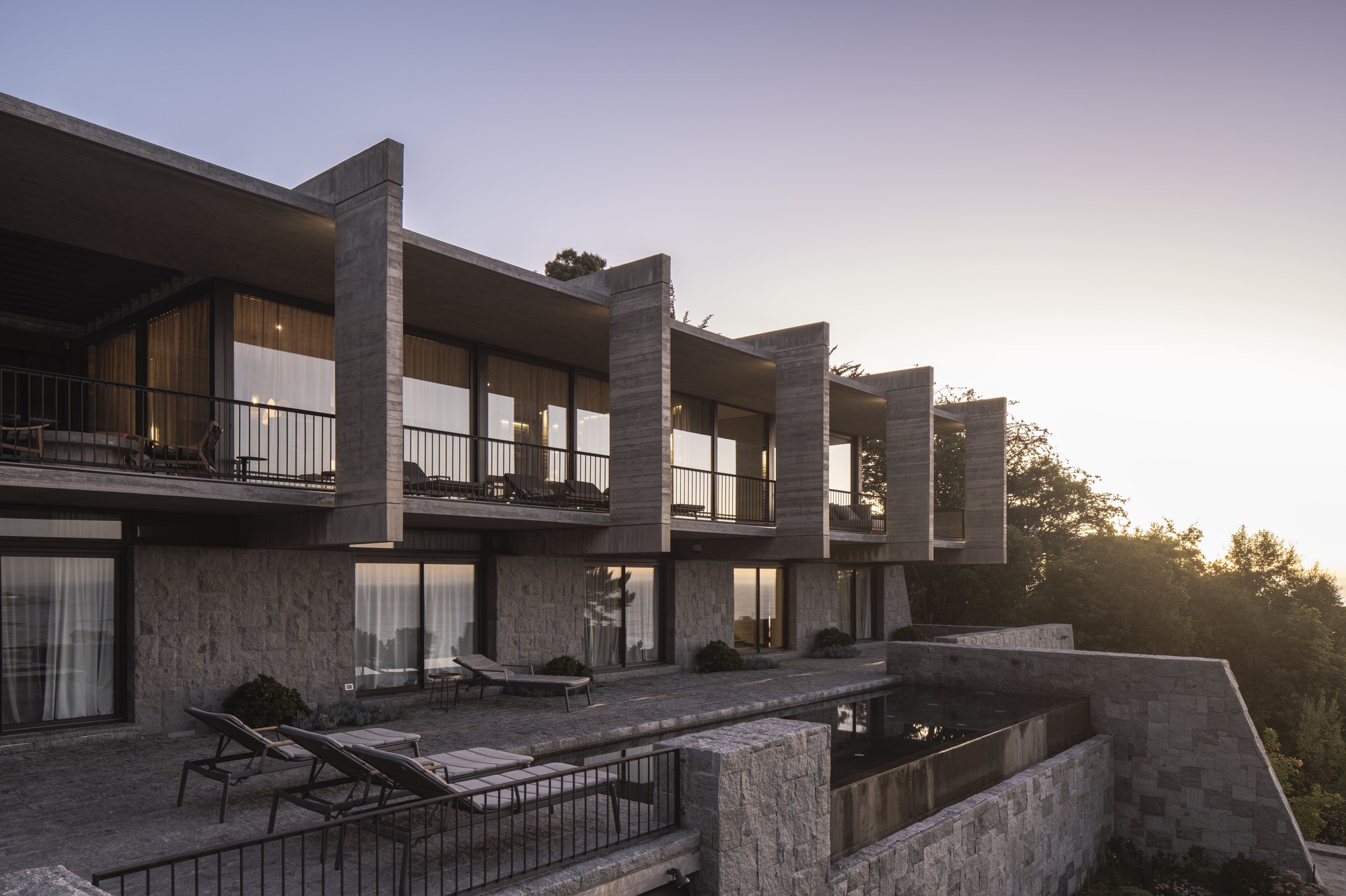 Stepped row of concrete fins and balconies above a stone terrace with sun loungers and a reflecting pool at sunset