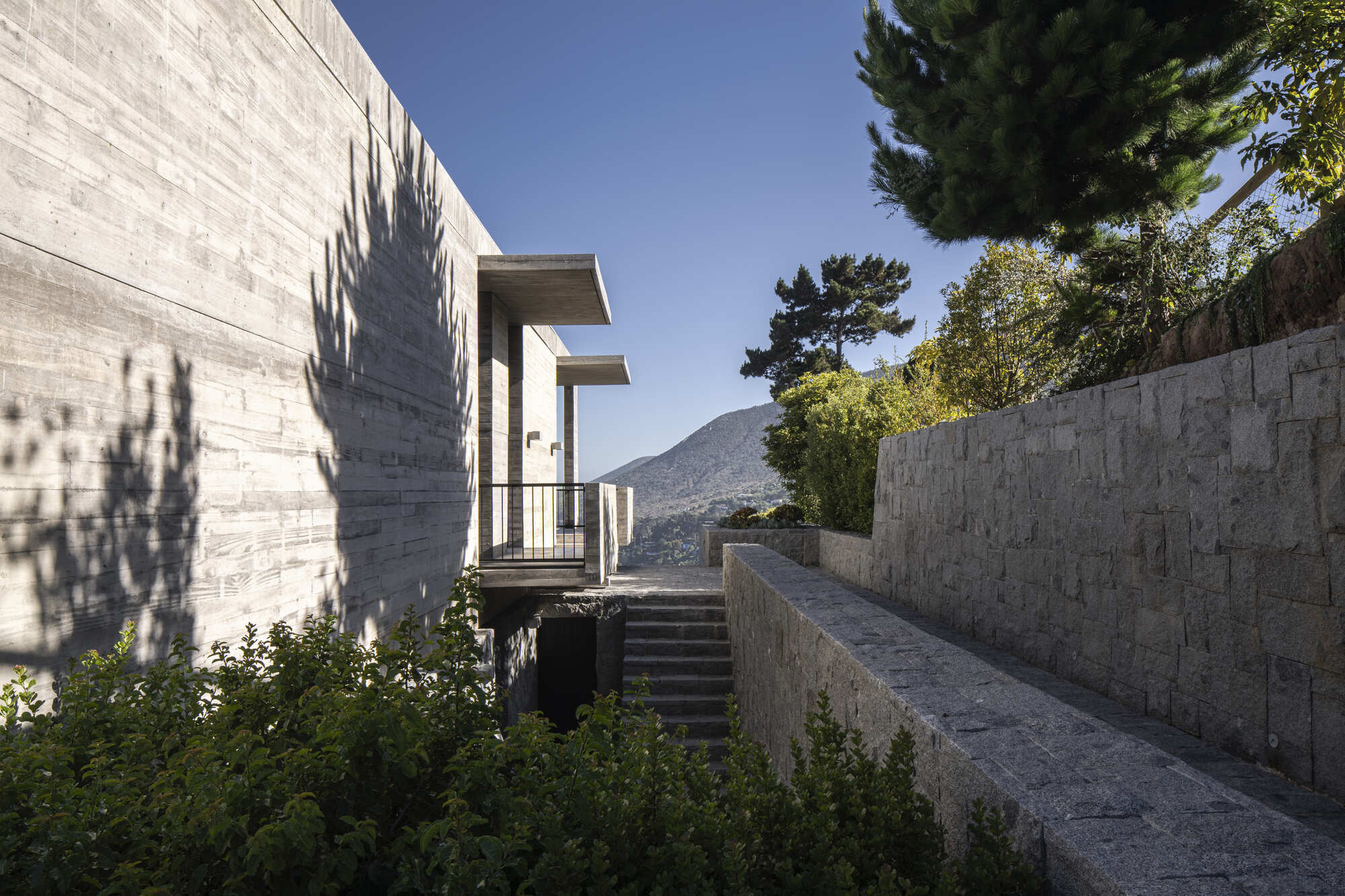 Narrow granite-walled passage with stone steps leading toward a cantilevered concrete volume, tree shadows falling across the wall in afternoon sun