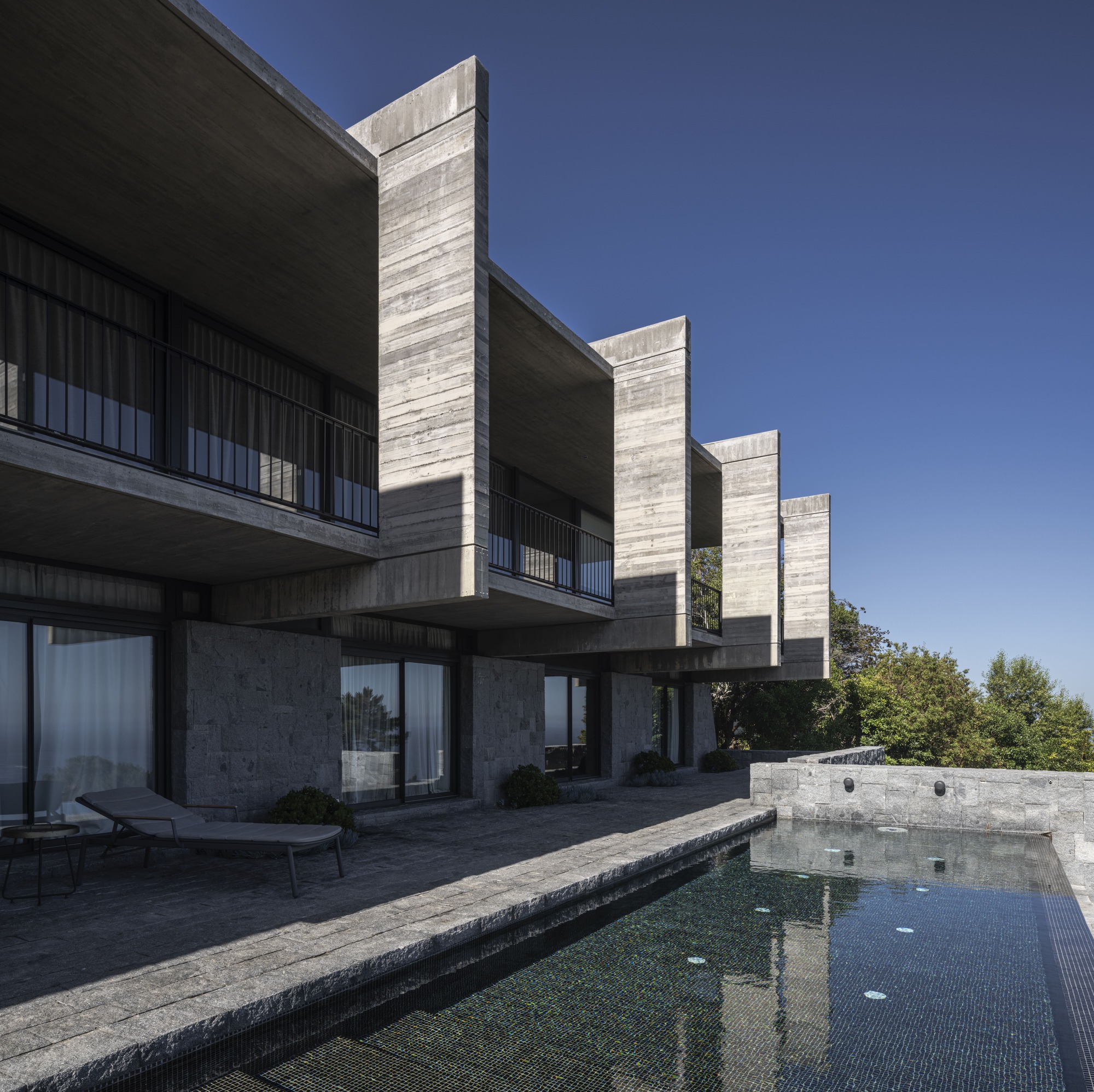 Row of projecting concrete fins and balconies above a stone-clad ground floor and dark tiled pool, seen in clear sunlight