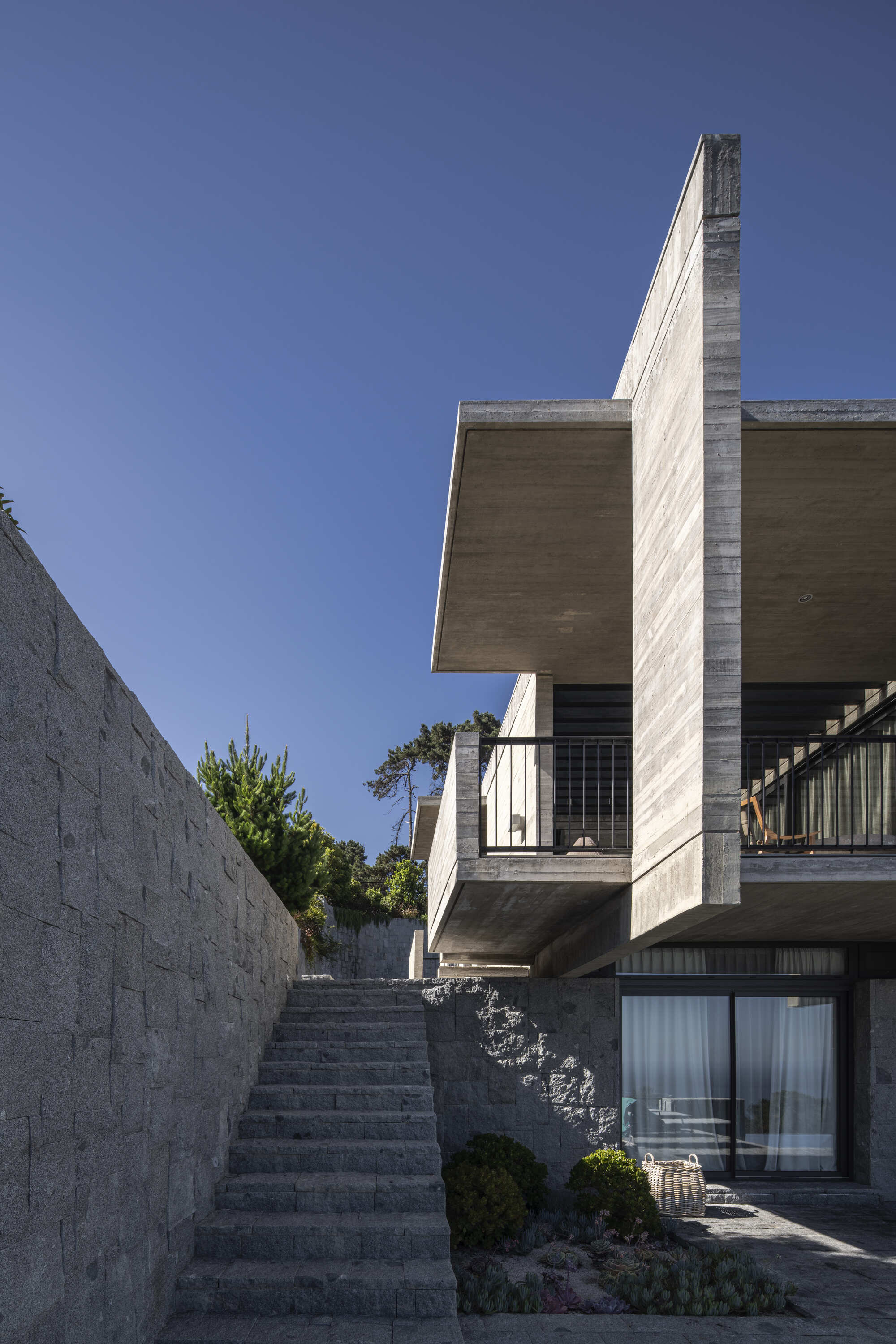 Stone stair climbing beside a retaining wall toward a cantilevered concrete slab and balcony with black railings under a clear sky
