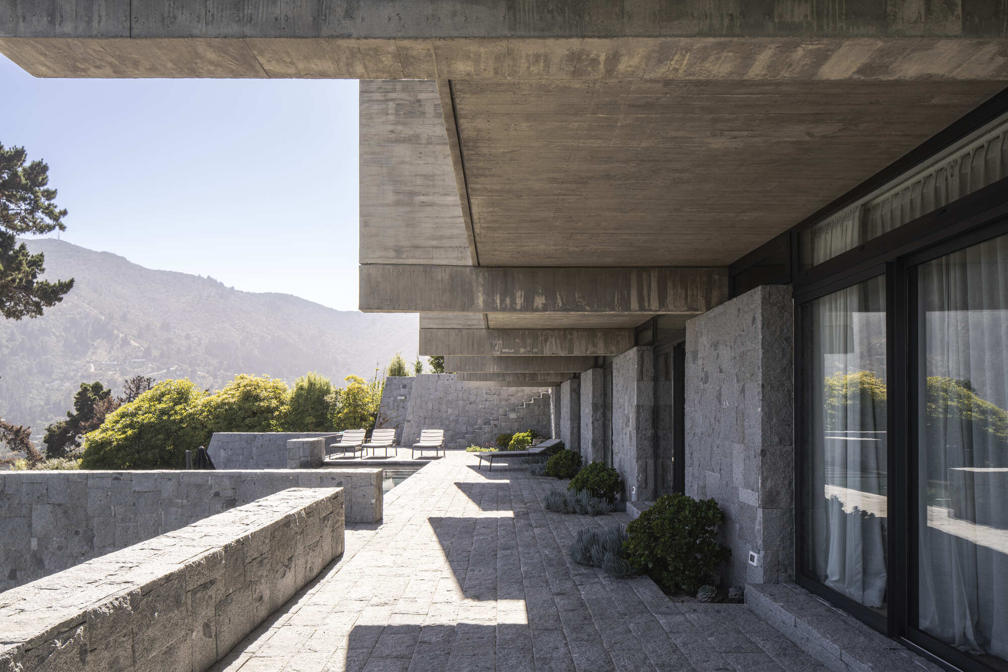 Covered terrace under stacked concrete beams with granite paving, stone columns and sliding glass doors, mountains hazy in the distance