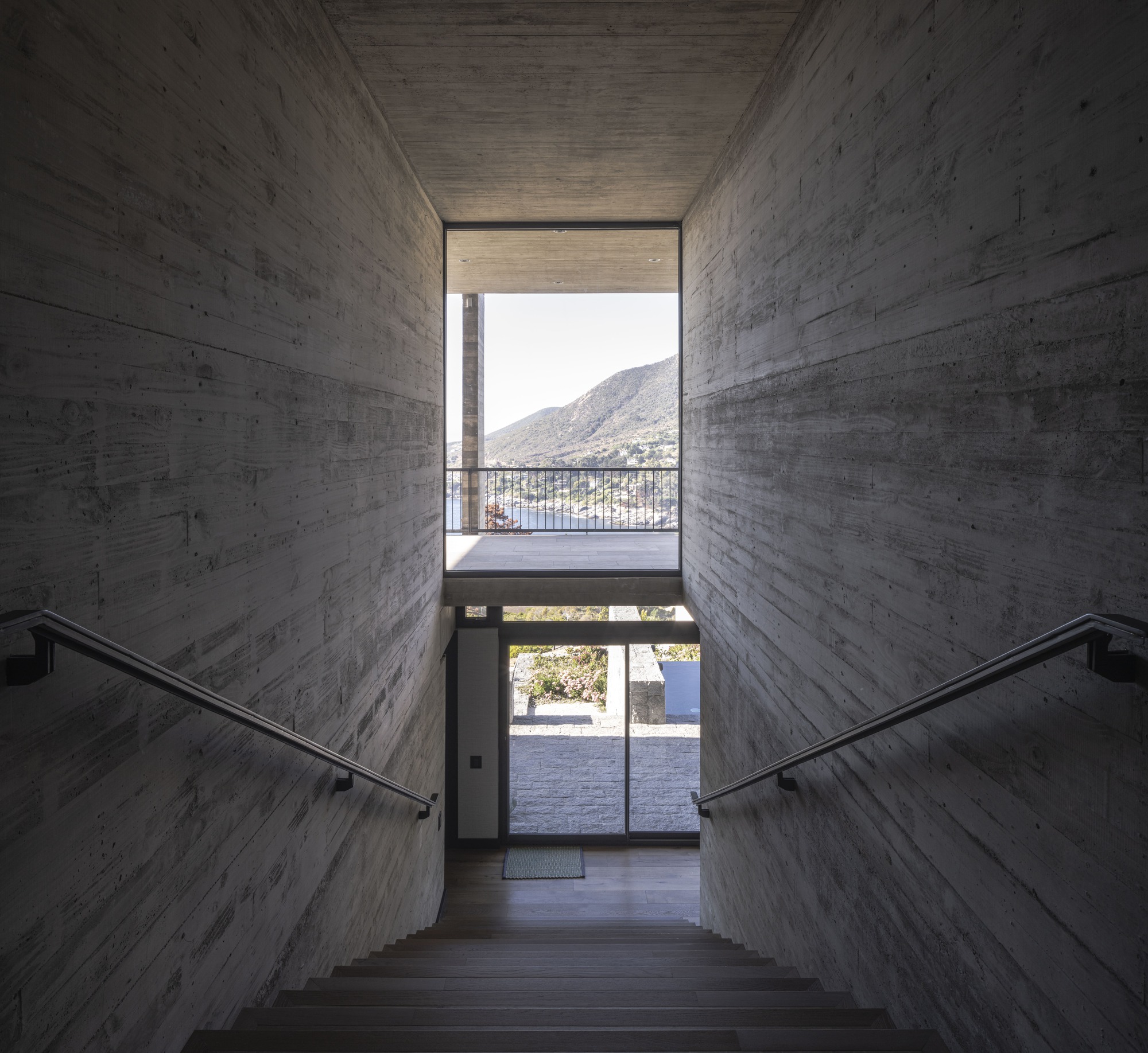 Concrete stairwell with steel handrails framing a view of a balcony, hillside and water beyond in bright daylight