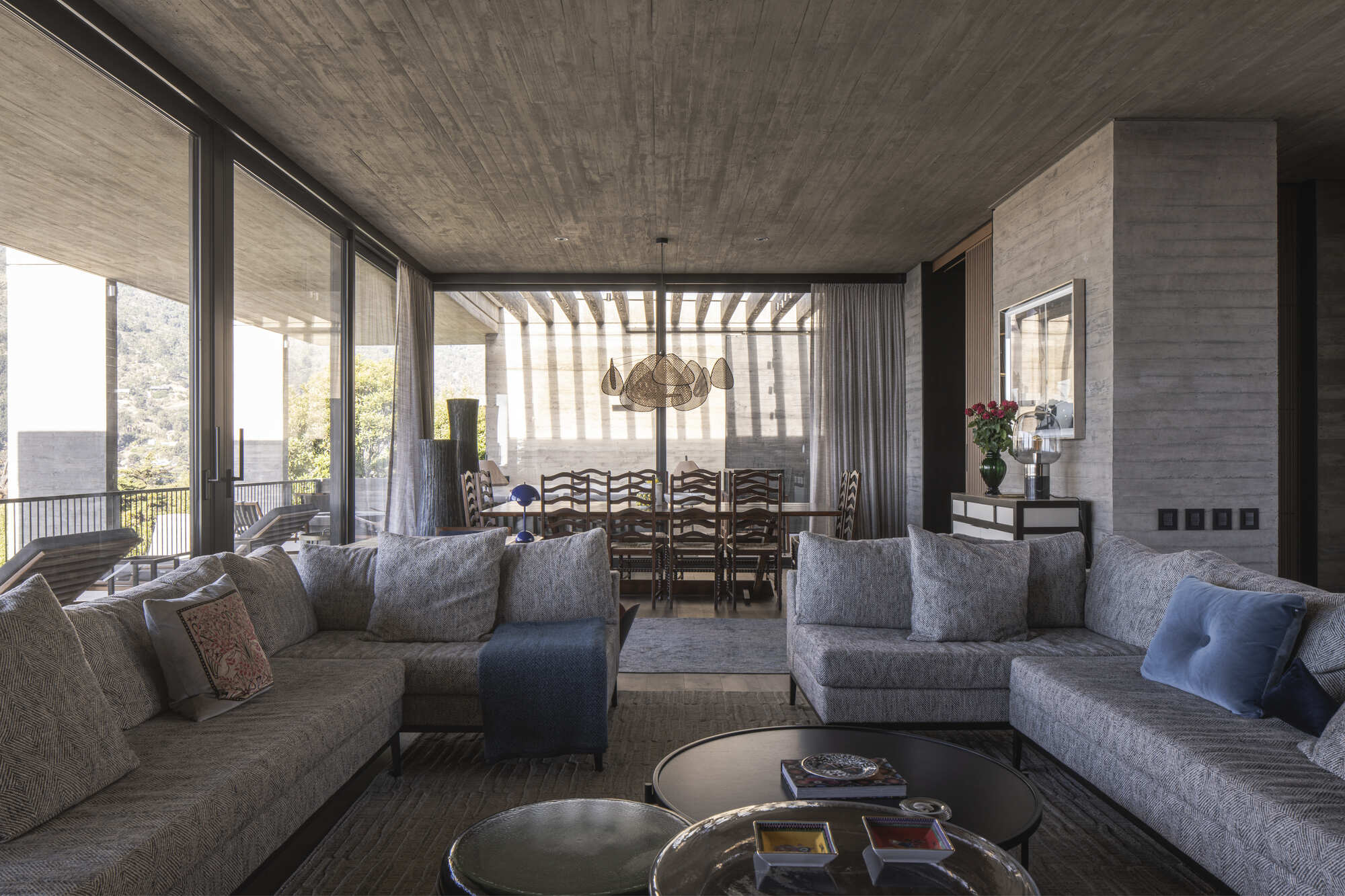 Living room with gray upholstered sofas, round glass tables and board-marked concrete ceiling, opening through glazing to a dining table on the terrace