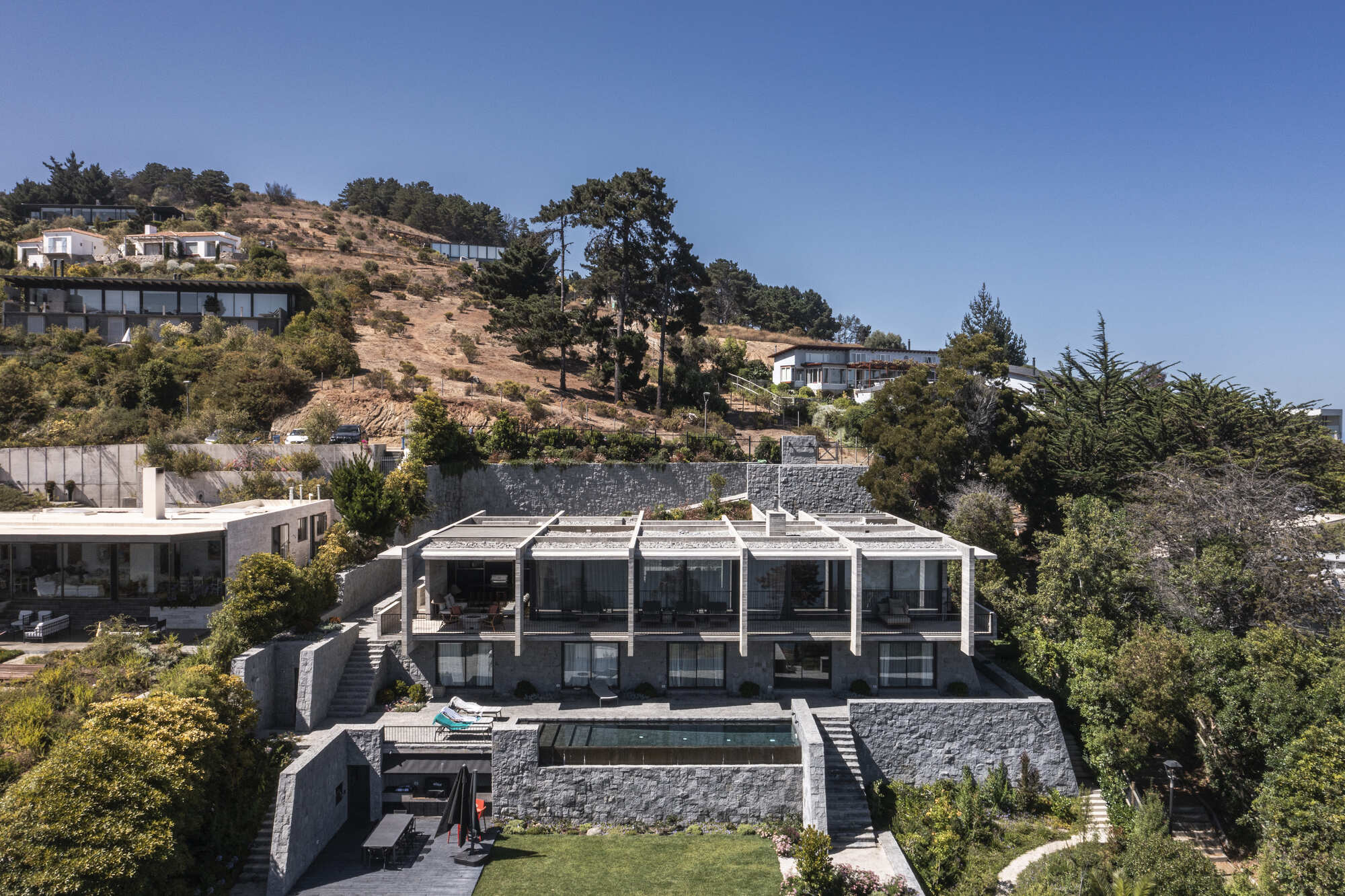 Drone view of the stepped house with pergola roof and stone retaining walls, a pool and lawn below, on a sunny hillside