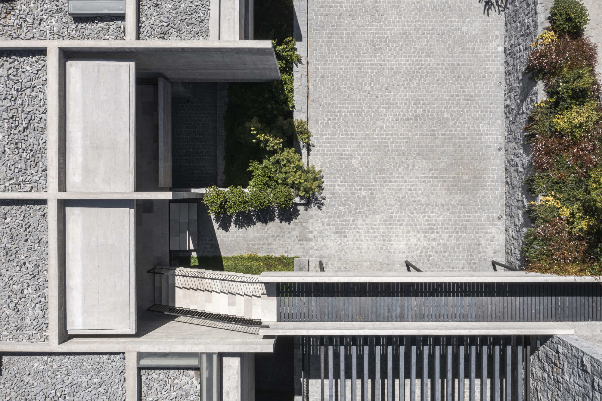 Top-down view of concrete frame, cobbled courtyard, stone-clad walls and a slatted bridge, with planted beds casting shadows in sunlight