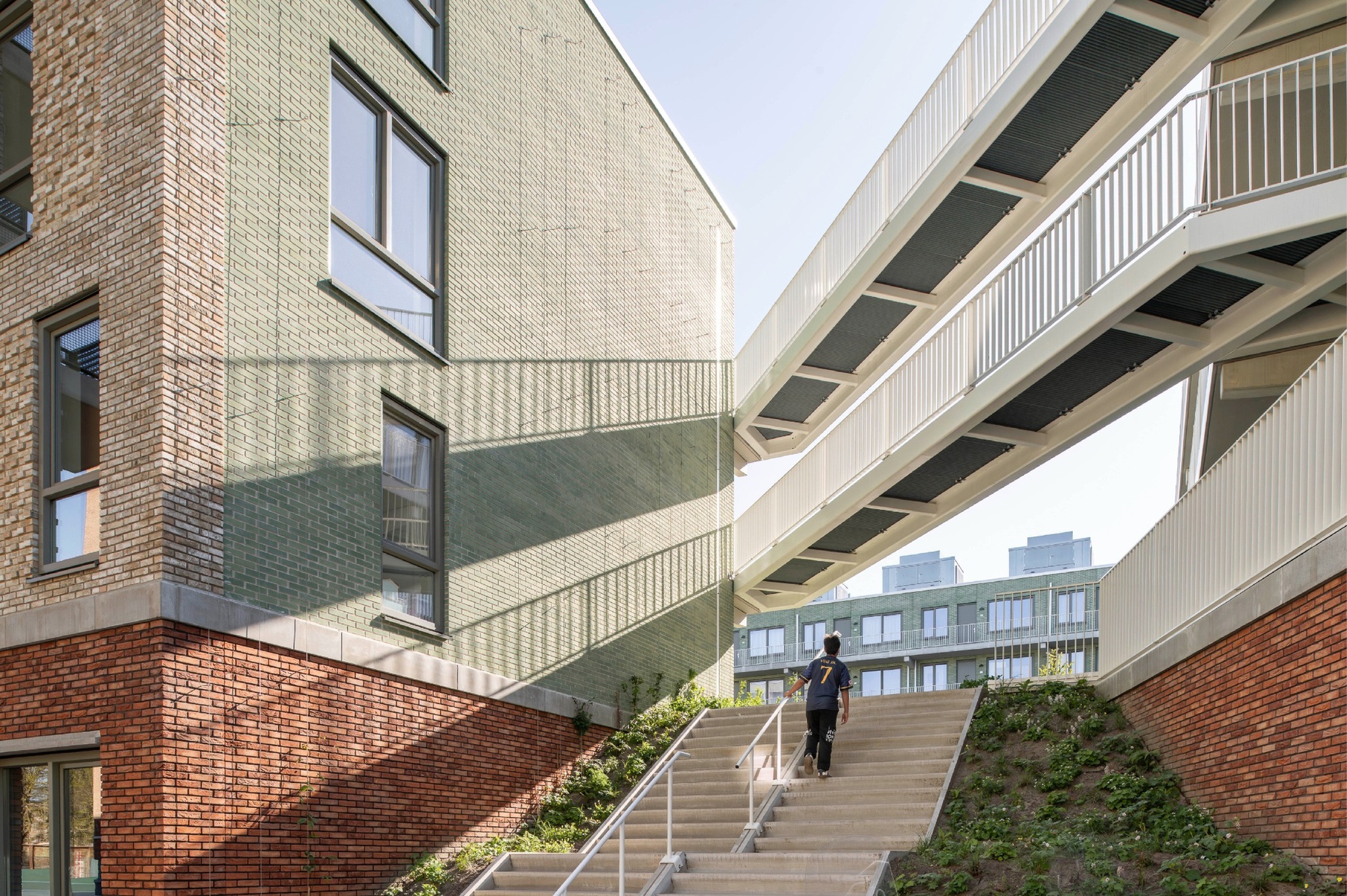 Concrete stair climbing between a green glazed brick gable and white steel walkways, with a person ascending in bright sun