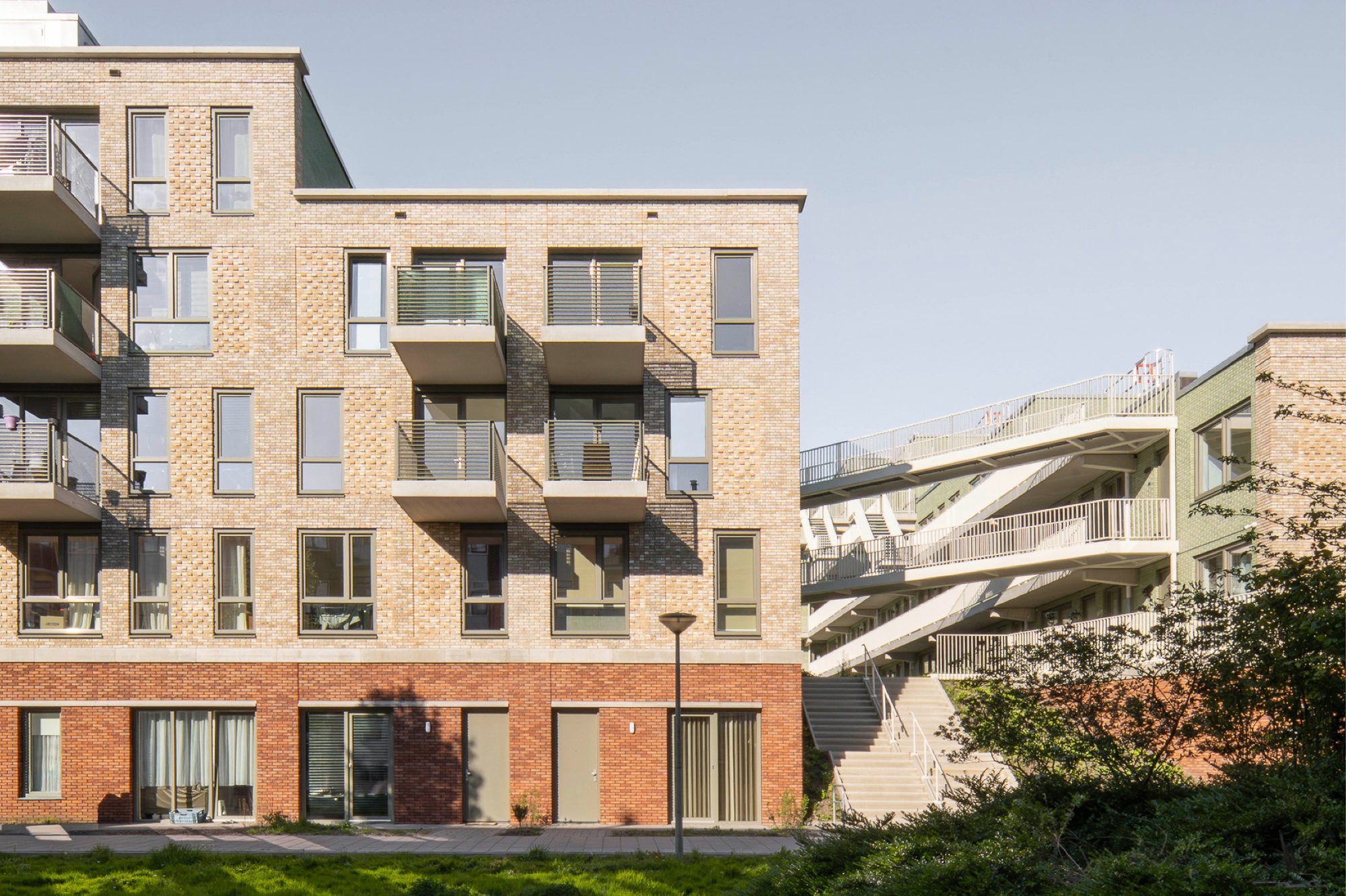Light brick apartment block with balconies beside external white stairs and walkways, seen across a lawn in low sun