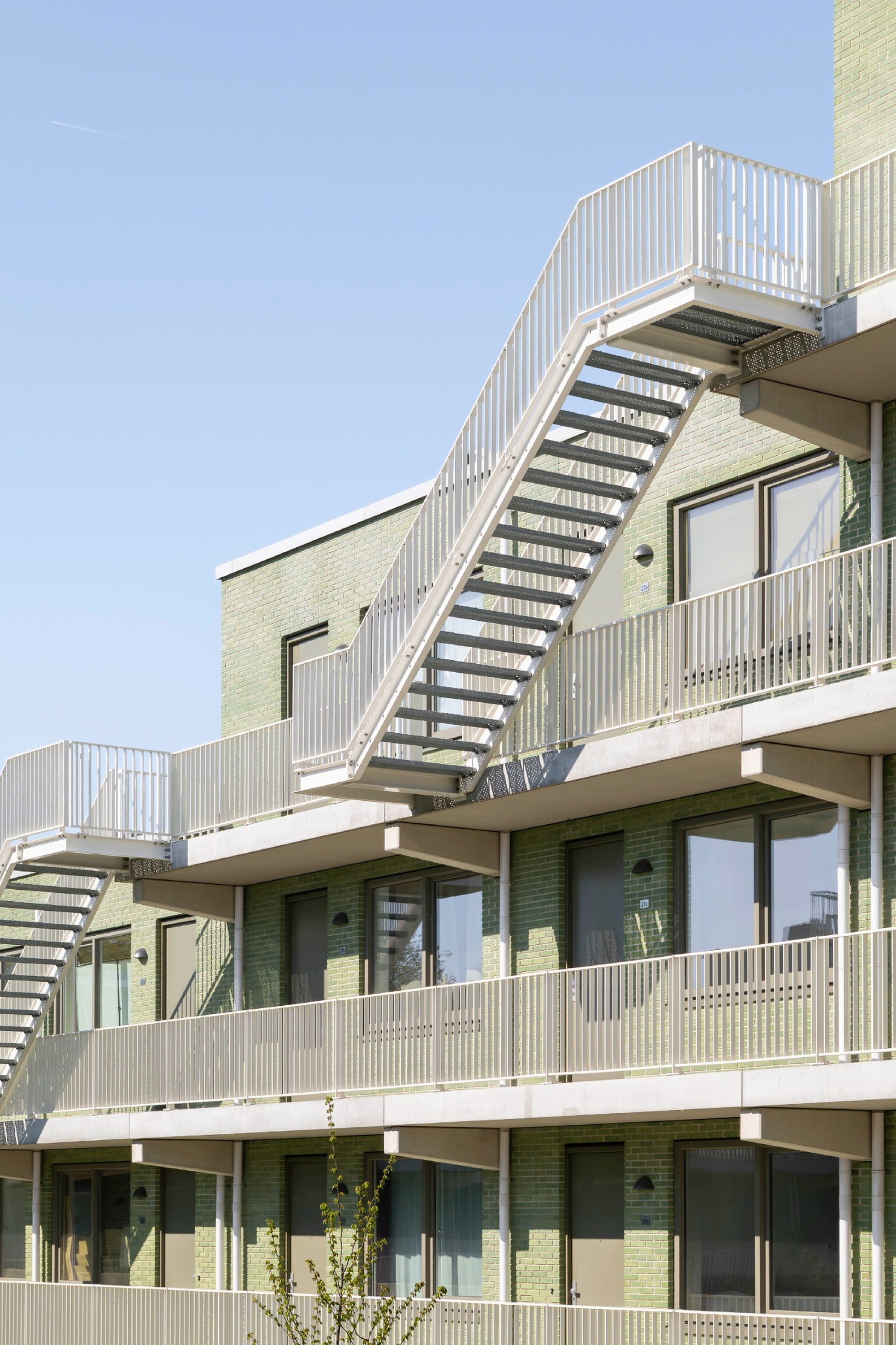 Green brick facade with white steel balcony railings and a diagonal open-tread stair under a clear blue sky