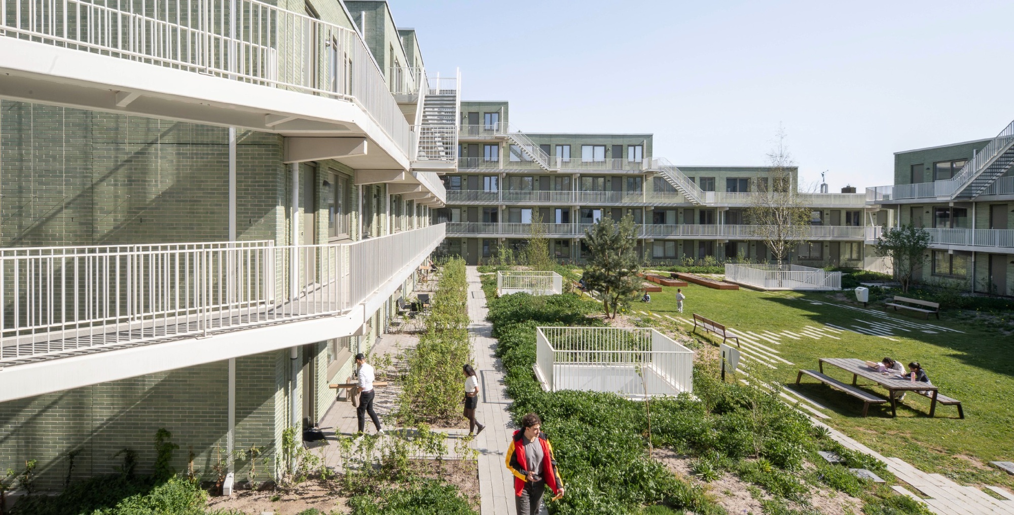 Courtyard garden between green brick blocks with white access galleries, planted beds and people sitting at a picnic table