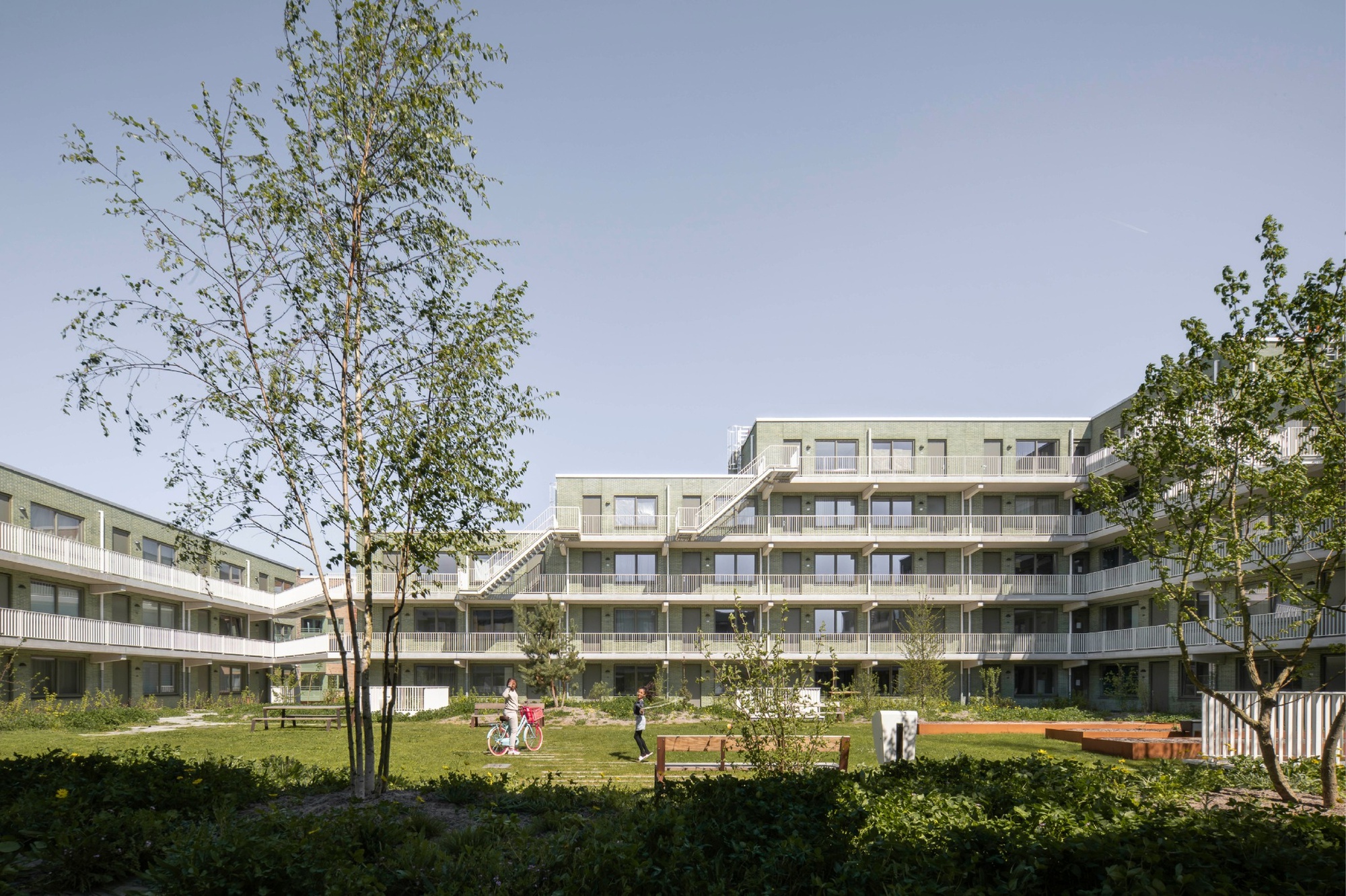 Terraced green brick courtyard block with white galleries seen through birch trees, a person pushing a bicycle across the lawn