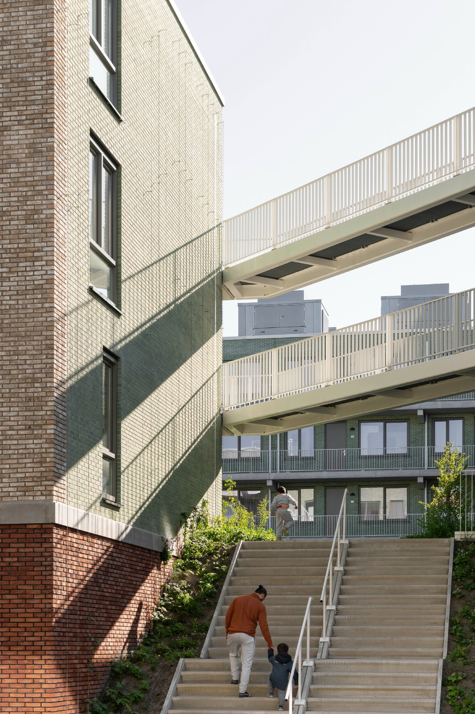 Adult and child climbing a wide concrete stair beneath white walkway bridges, with shadows falling across the green brick wall