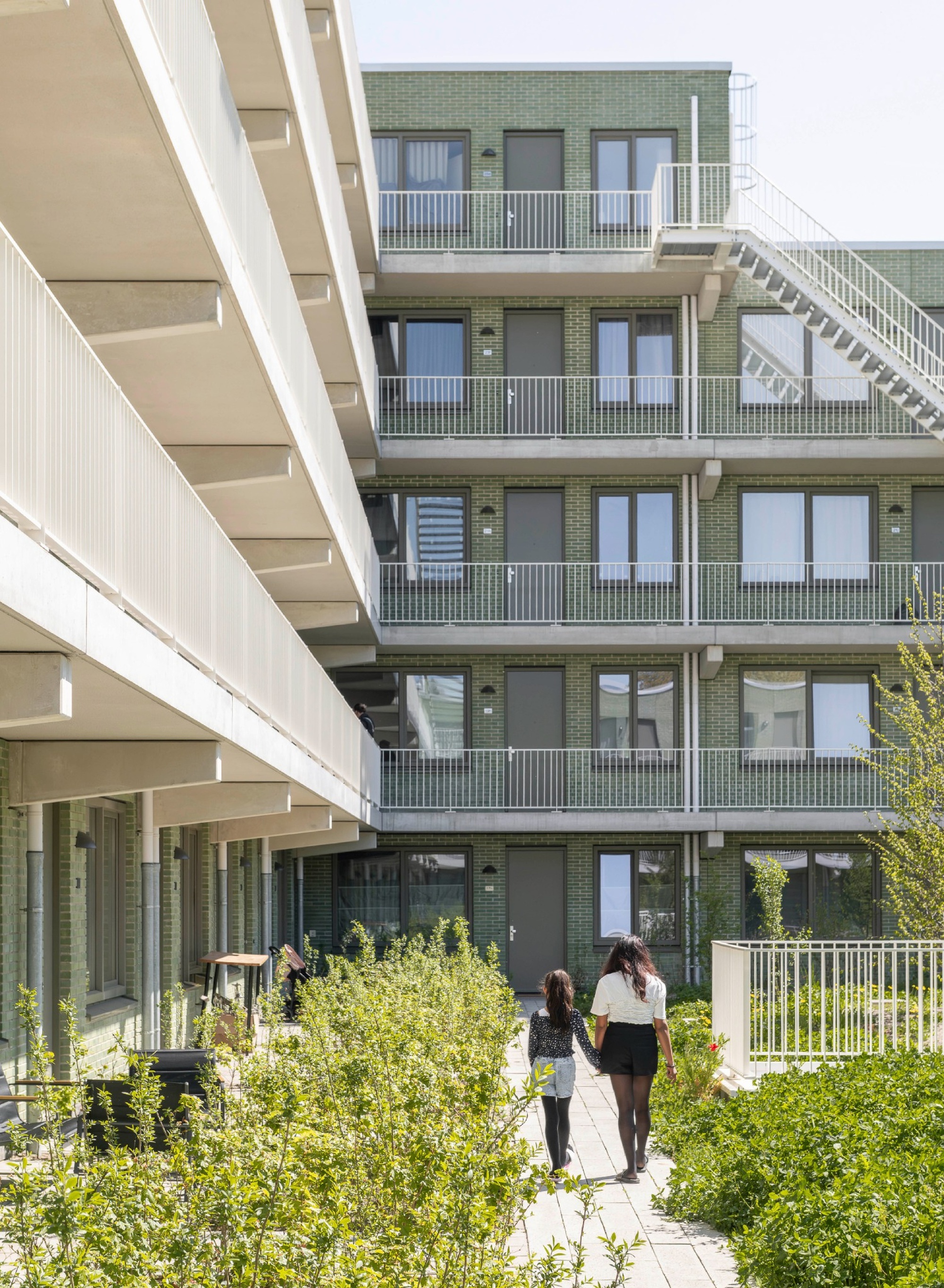 Green brick courtyard facade with white balconies and an exterior steel stair, a woman and child walking the paved path through planting