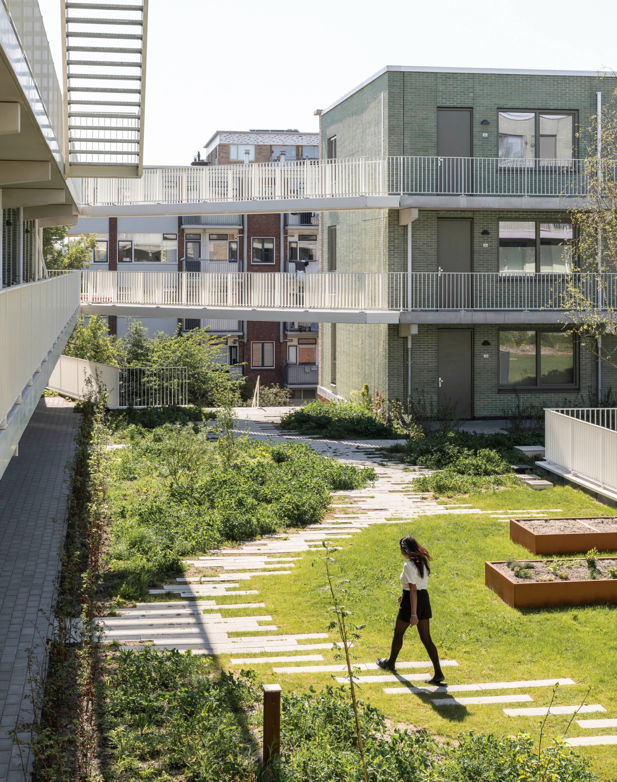 Courtyard lawn crossed by a staggered stepping-stone path, with a woman walking and corten planter beds beside green brick walls