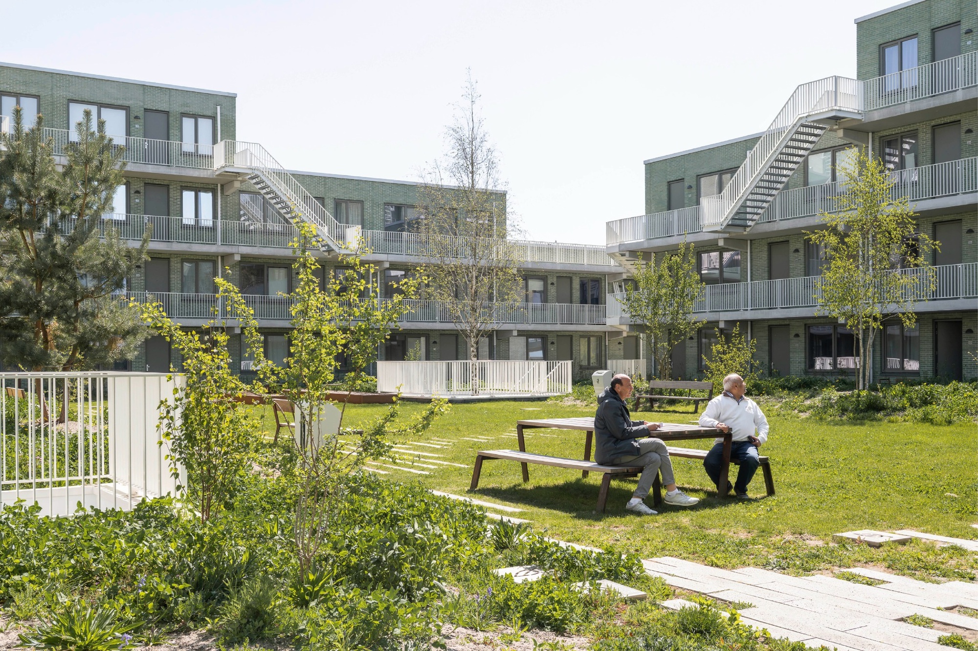 Two men talking at a picnic table on the courtyard lawn, surrounded by young trees and balcony-access apartment blocks in sunshine