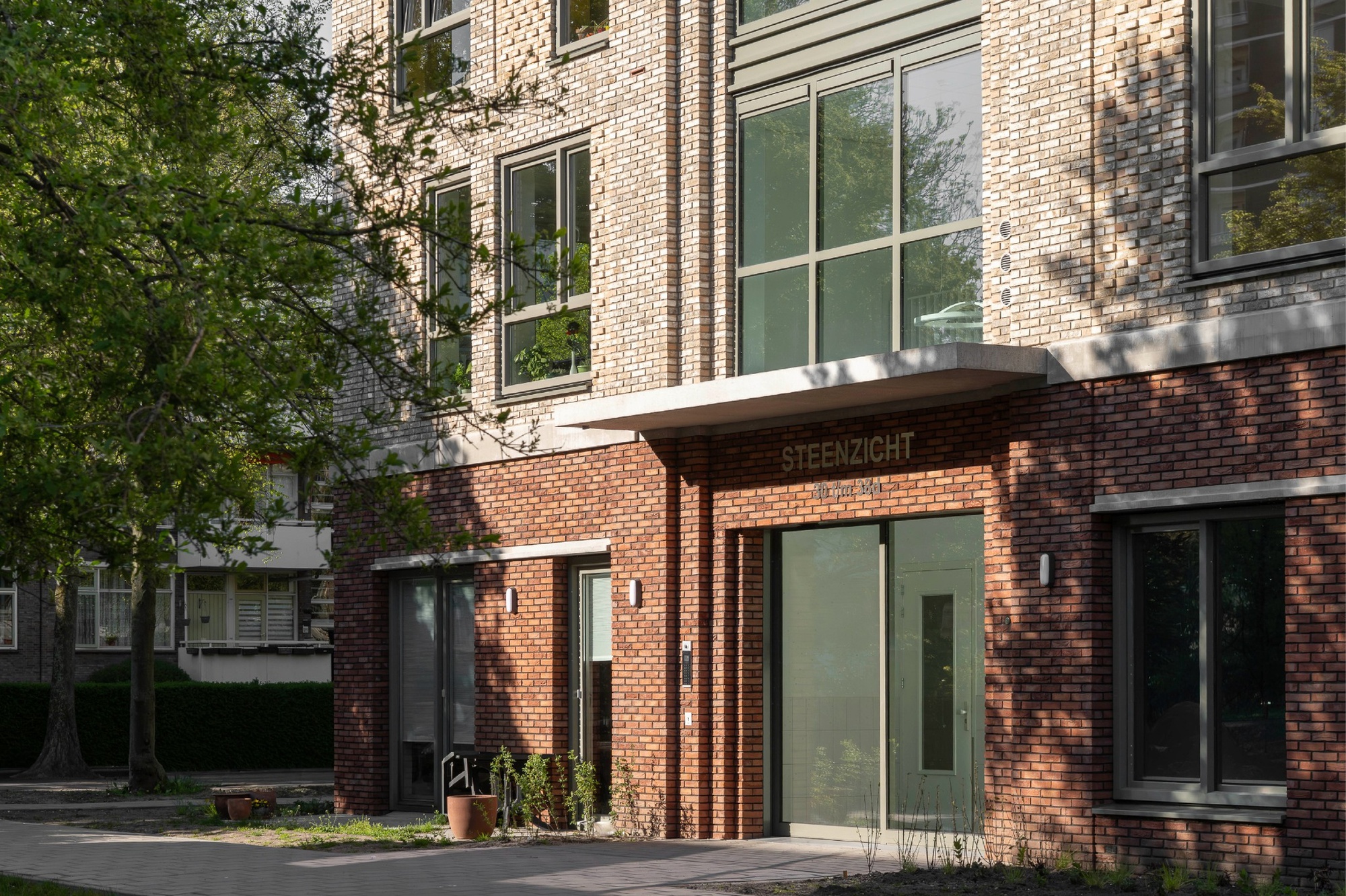 Close-up of the red brick entrance with a concrete canopy and lettering above glazed doors, dappled by tree shadows