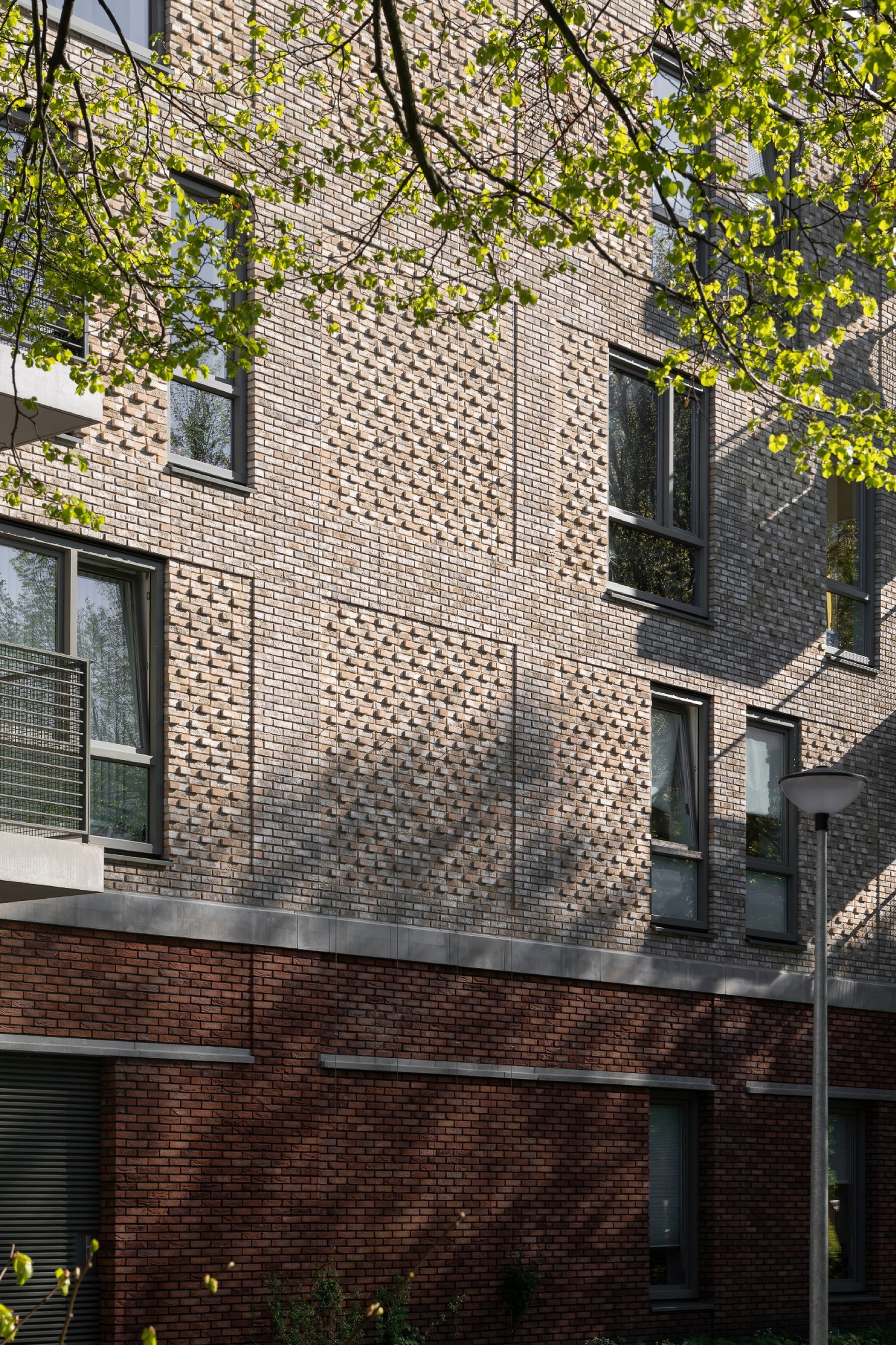 Patterned brick facade with projecting header bricks above a darker red brick base, dappled by spring leaves and shadow