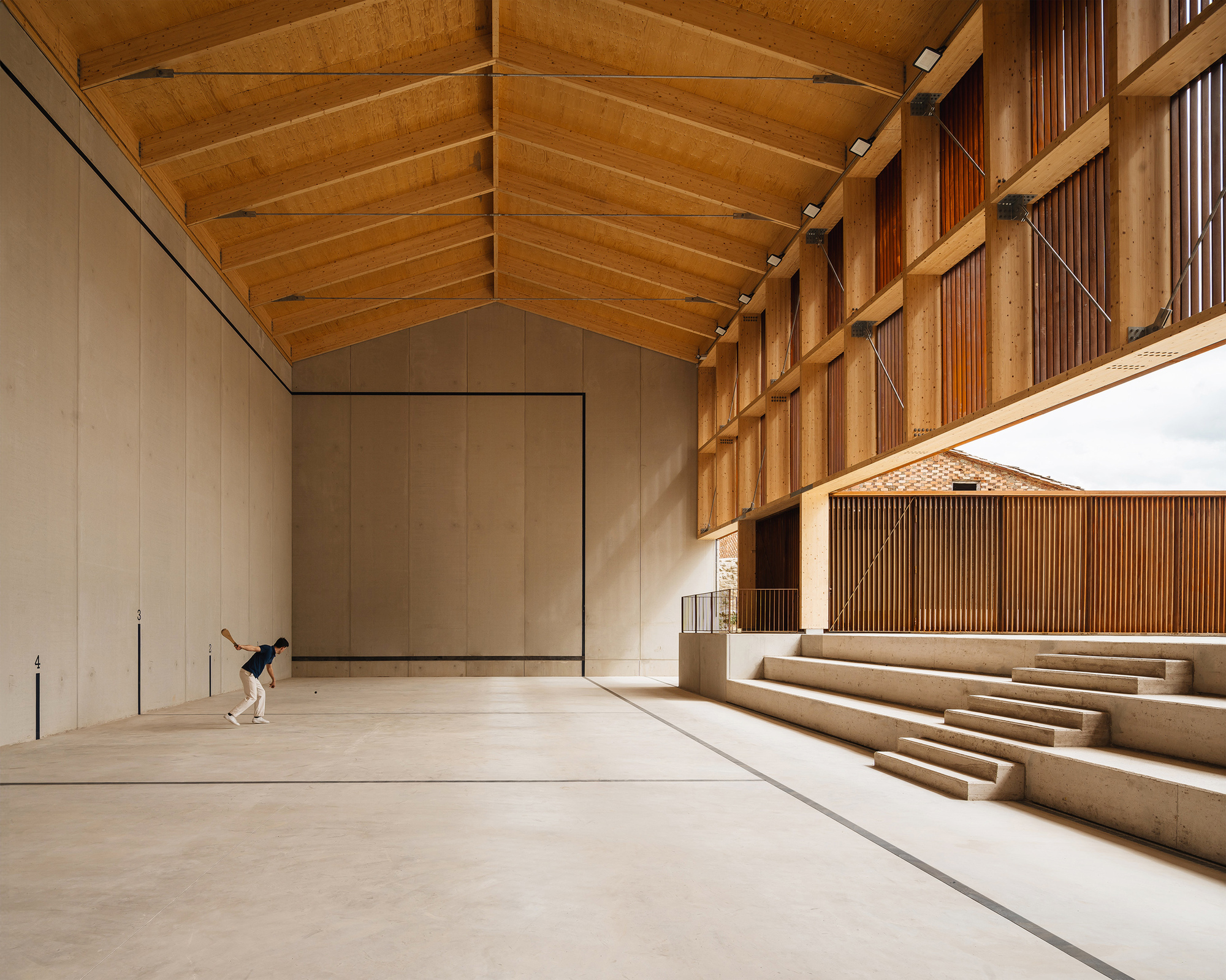 Concrete-walled pelota court with timber roof trusses, stepped seating and a lone player mid-swing on the pale floor
