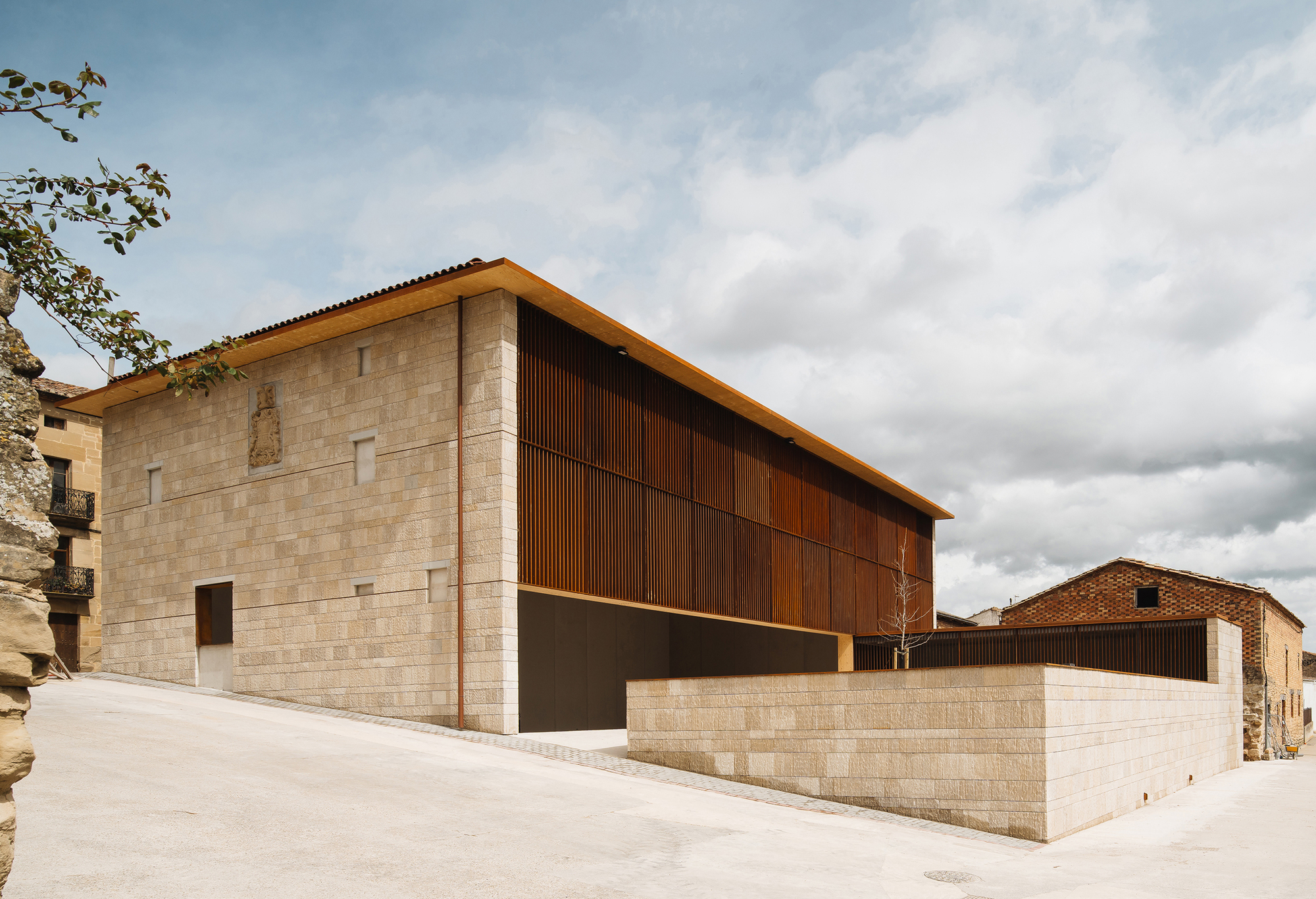 Stone-clad corner volume with a rust-toned slatted upper floor and projecting timber eave under a cloudy sky