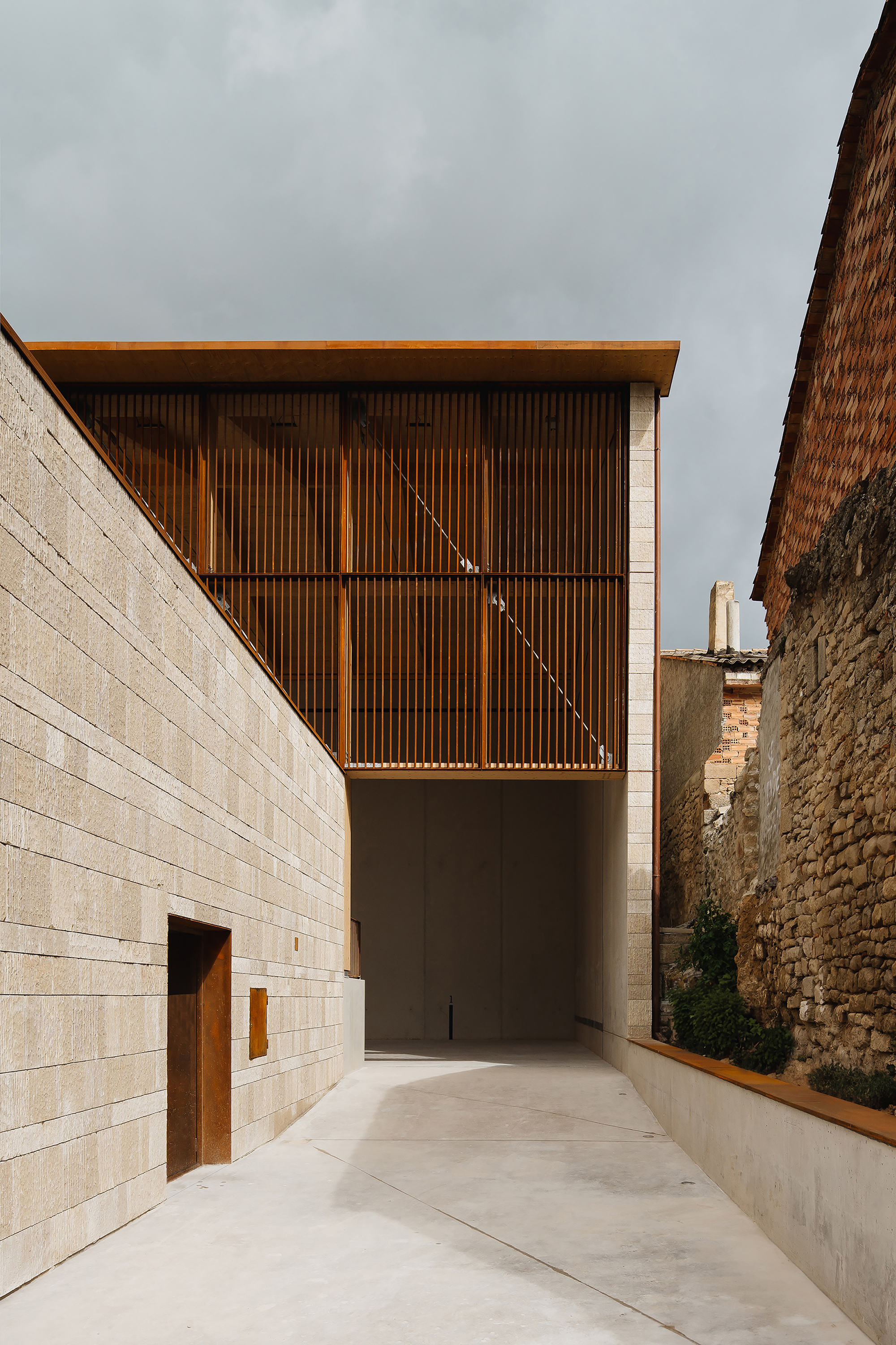 Narrow passage between a stone block wall and an old rubble wall, with a weathering steel slatted volume above