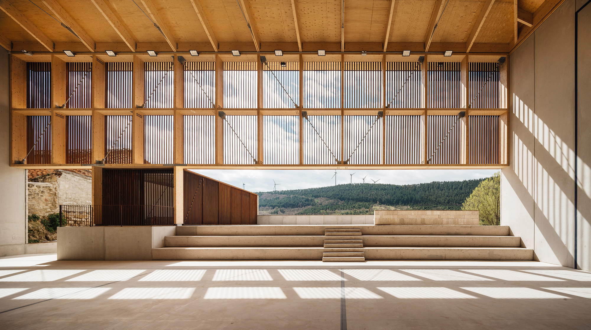Interior facing the open side of the hall, with sunlight casting slatted shadows across the floor and wind turbines on distant hills