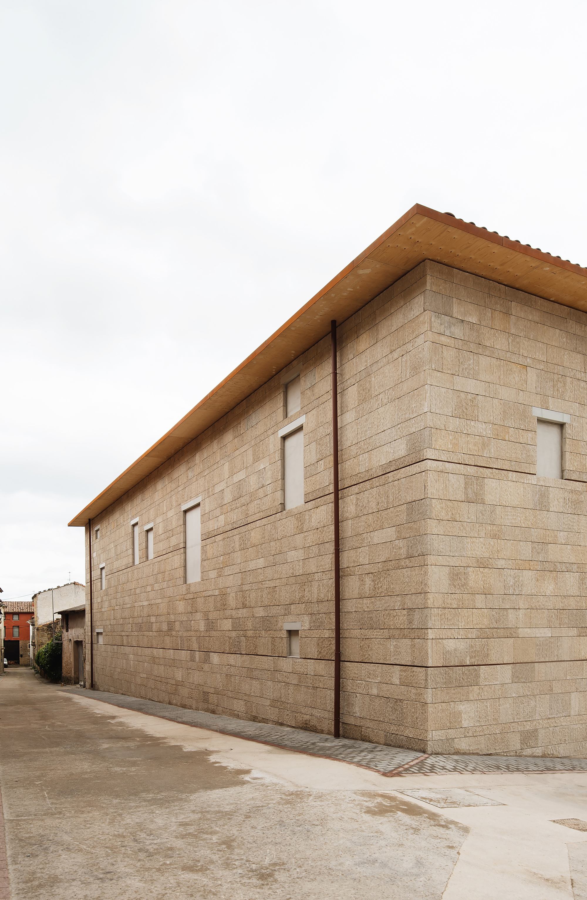 Long stone block side elevation with small recessed windows, a copper downpipe and a timber eave under an overcast sky
