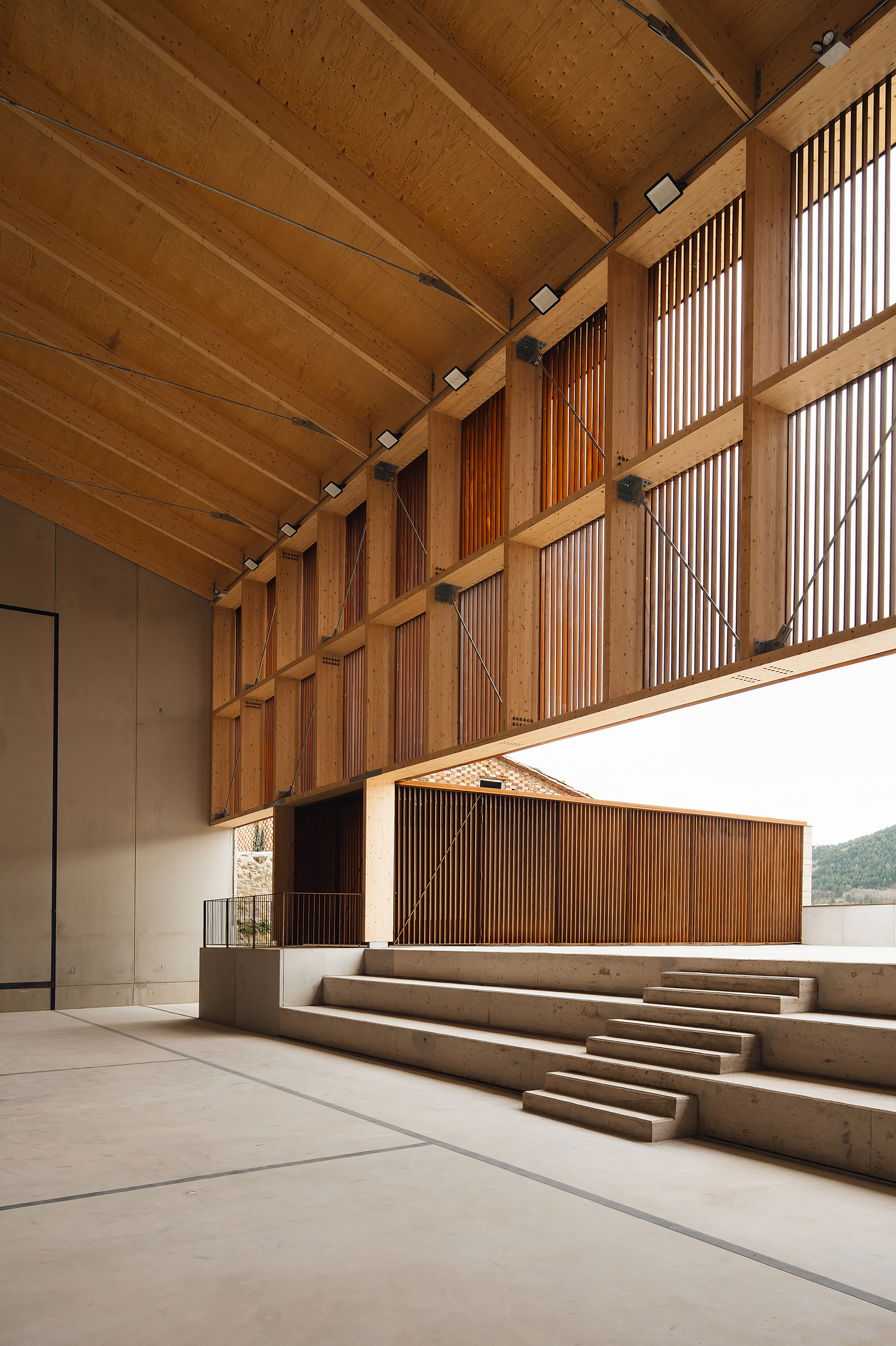 Low-angle view of glulam frame, angled tie rods and vertical timber slats above concrete bleacher steps in soft daylight