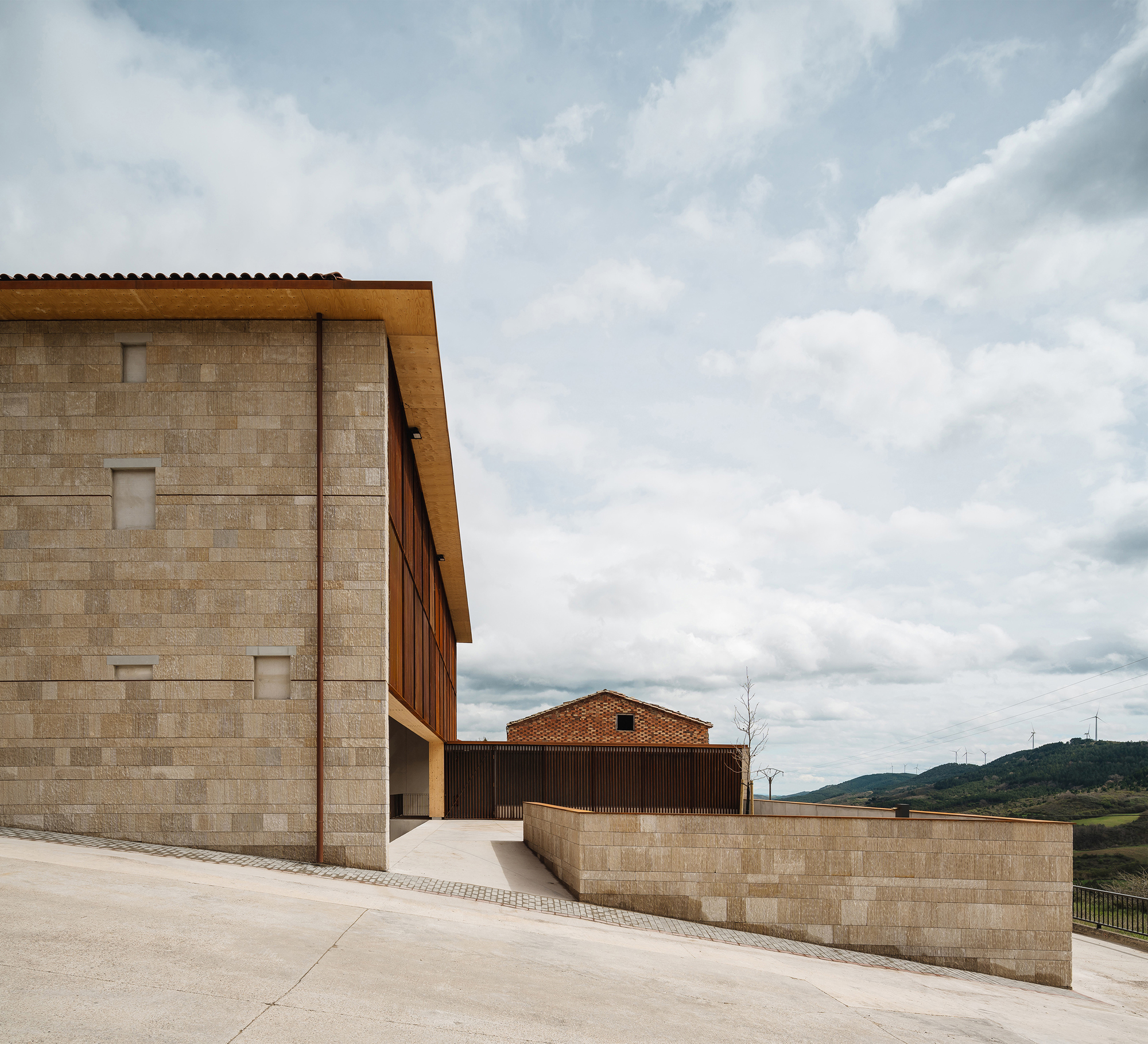 Stone block corner with a timber eave and slatted upper level beside a low retaining wall, under a broad cloudy sky