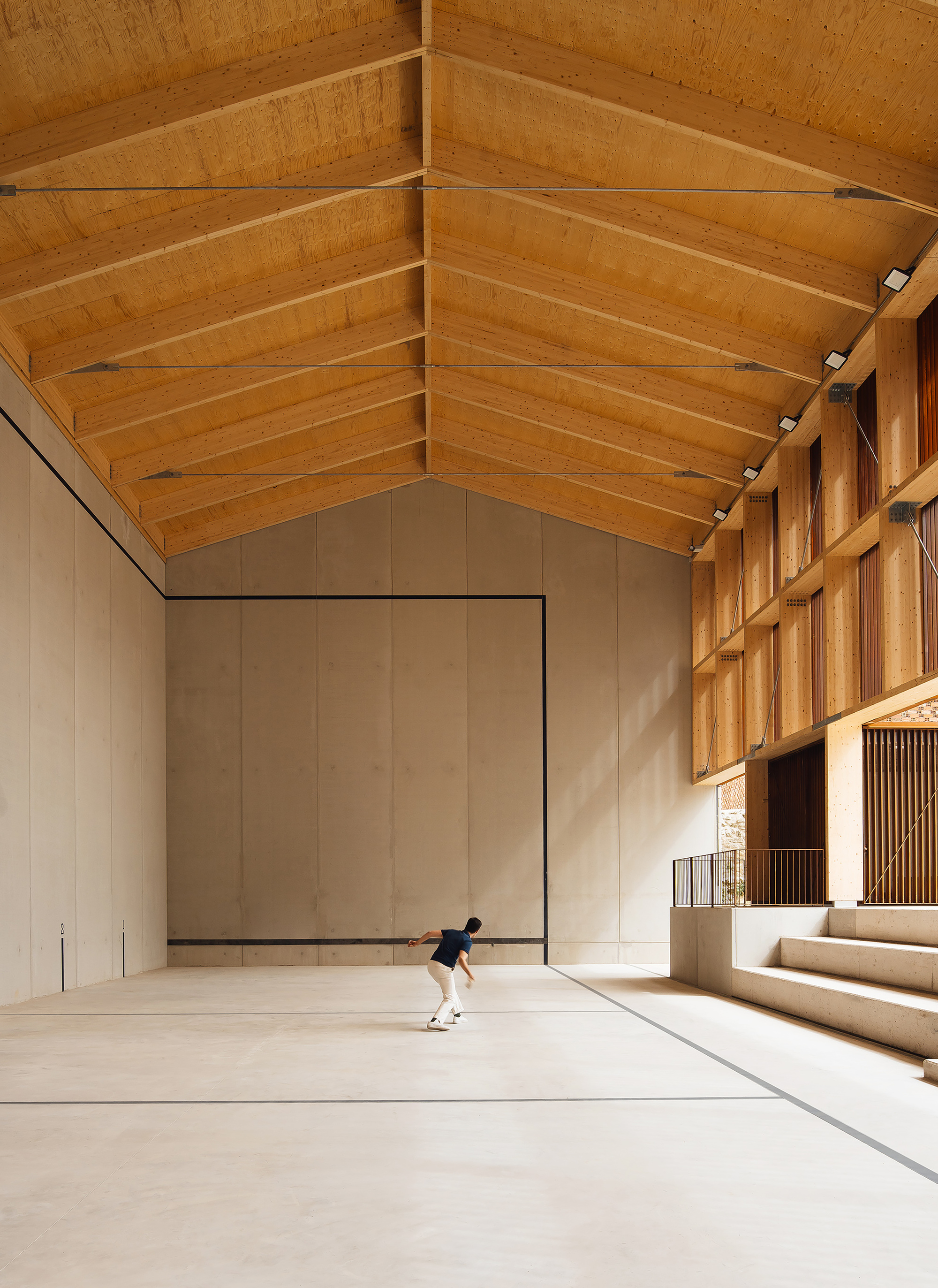 Interior of a pitched timber-roofed court with a concrete back wall, a lone player and stepped seating under warm light