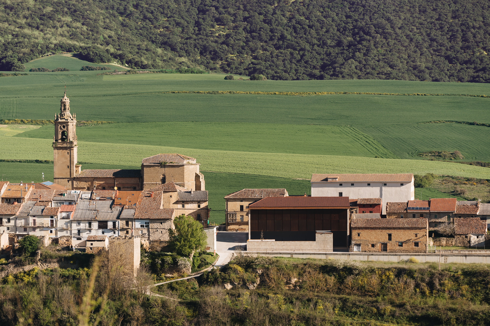 Distant view of the dark-clad building among tiled village roofs, a church bell tower and green fields on a sunny day