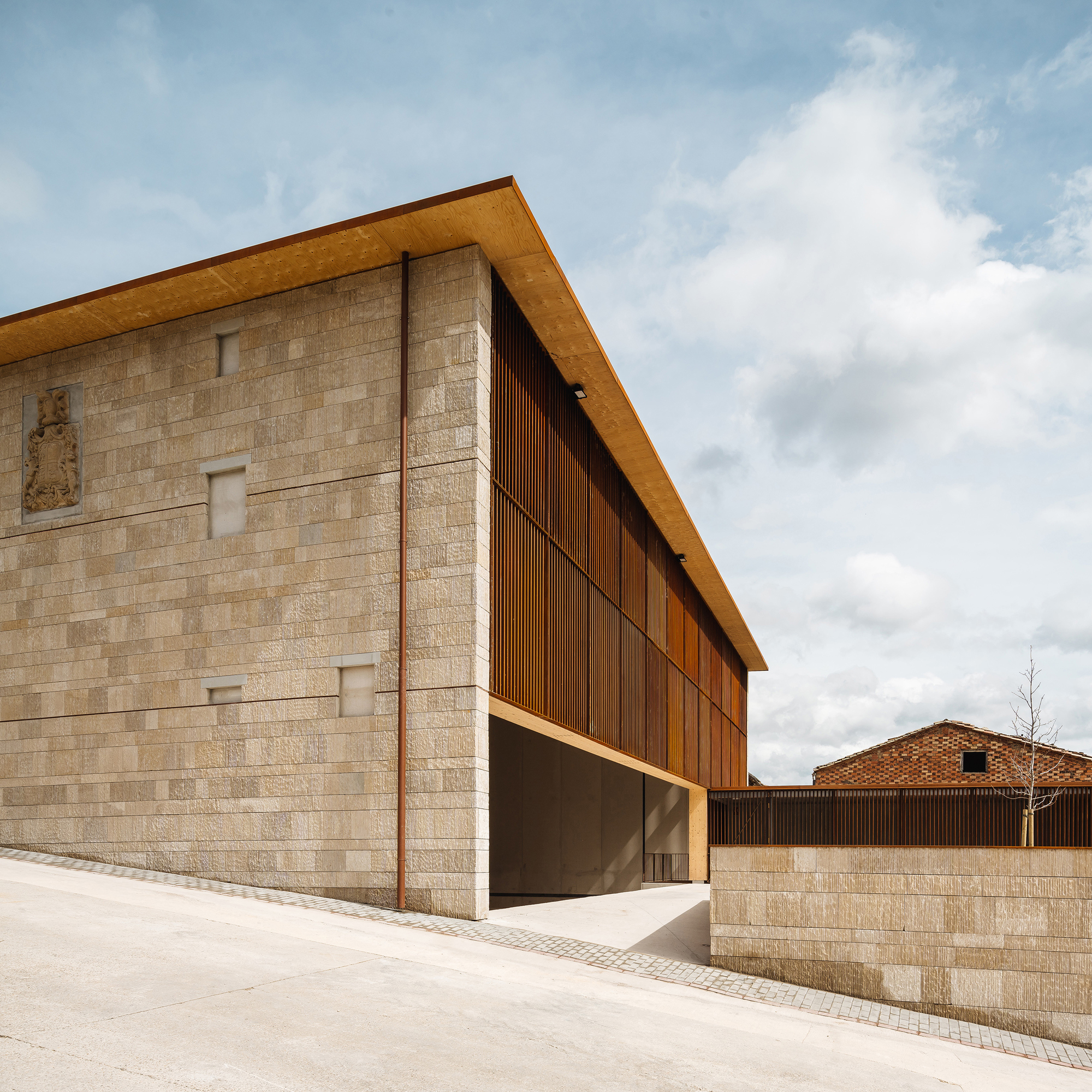Corner of the stone block volume with a carved coat of arms, timber soffit and slatted screen under clouds