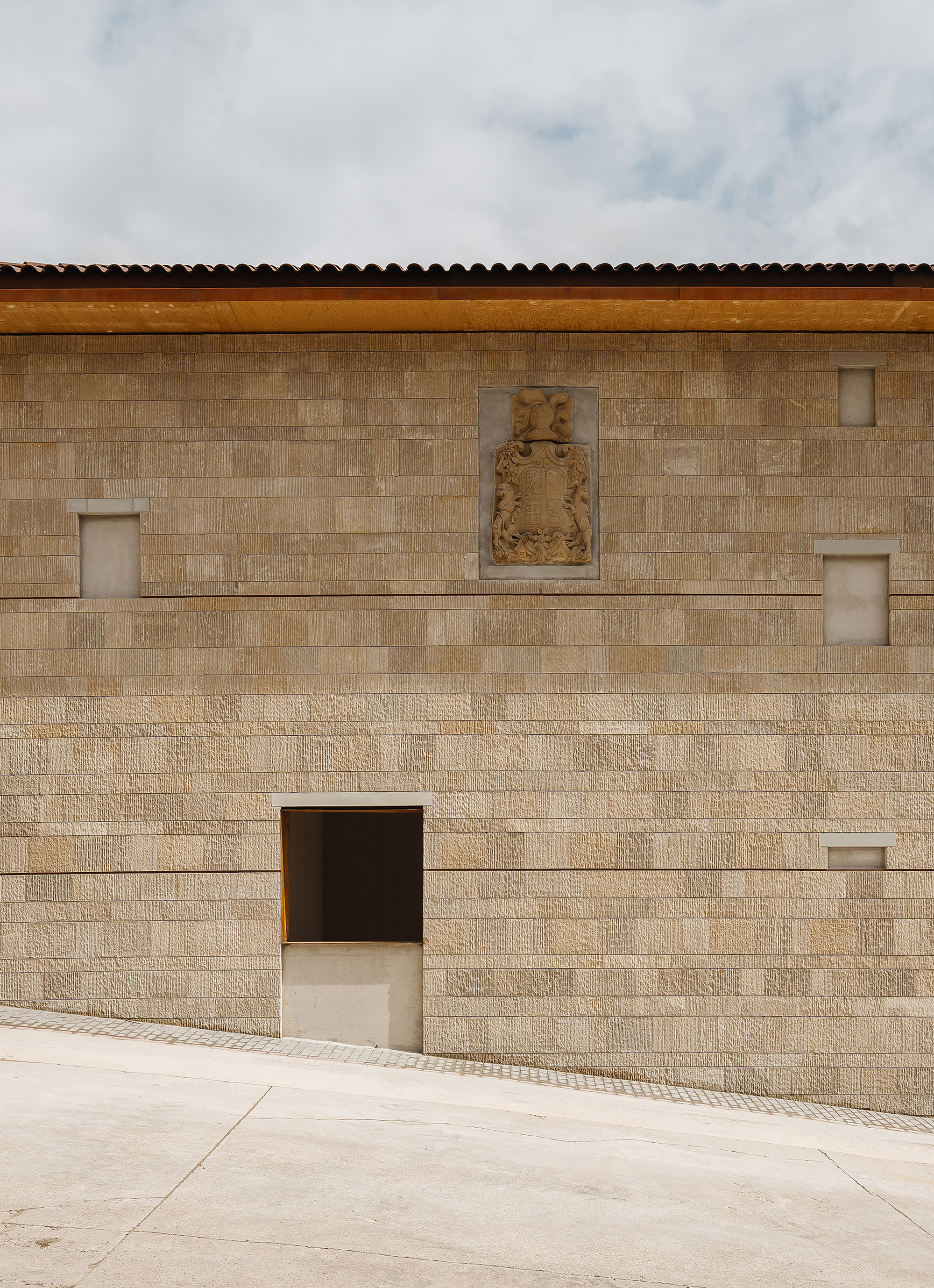 Stone block wall with a carved heraldic crest, small recessed windows and a timber fascia beneath a tiled eave