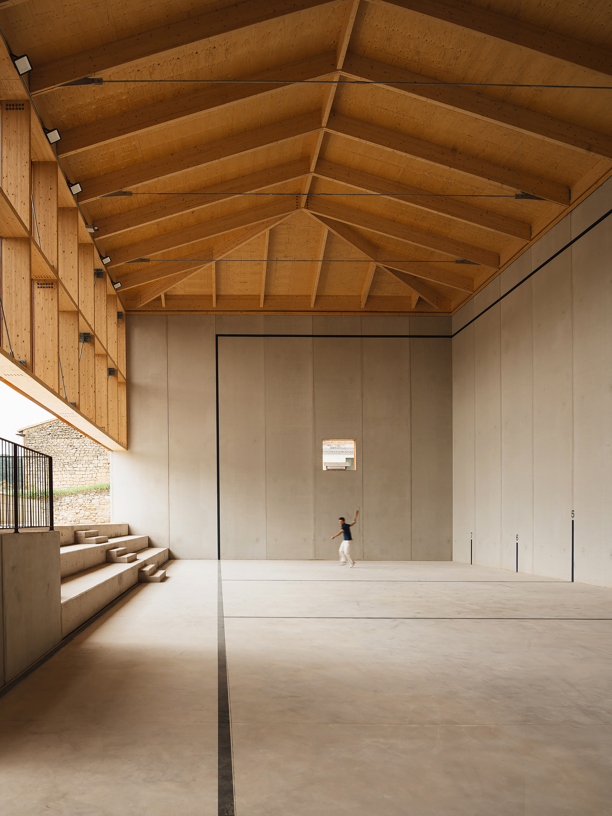 Court interior with glulam roof trusses, tiered concrete steps at left and a player mid-swing against the end wall