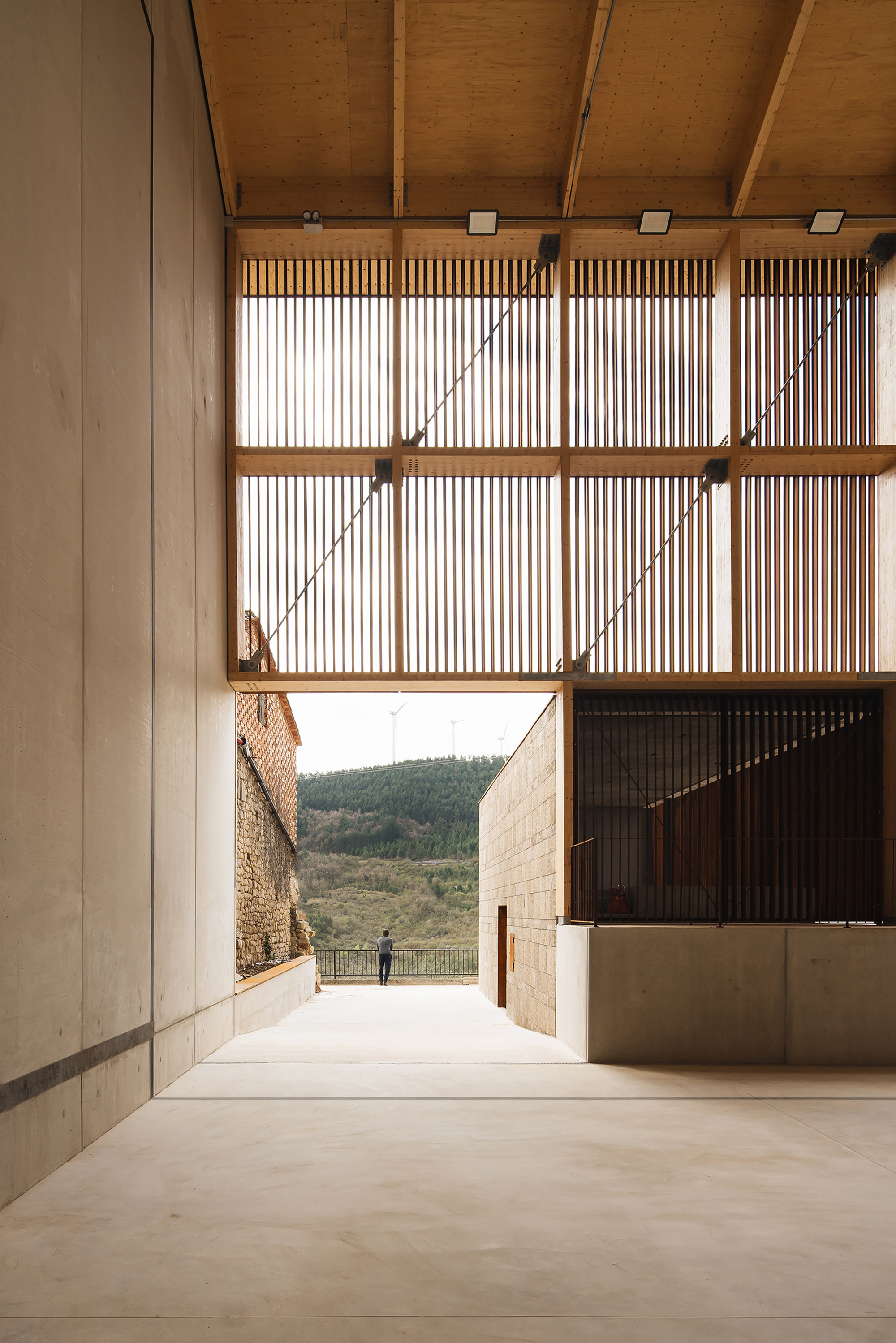 Timber slatted screen above a concrete passage framing a stone wall, wooded hillside and a person standing at the far railing