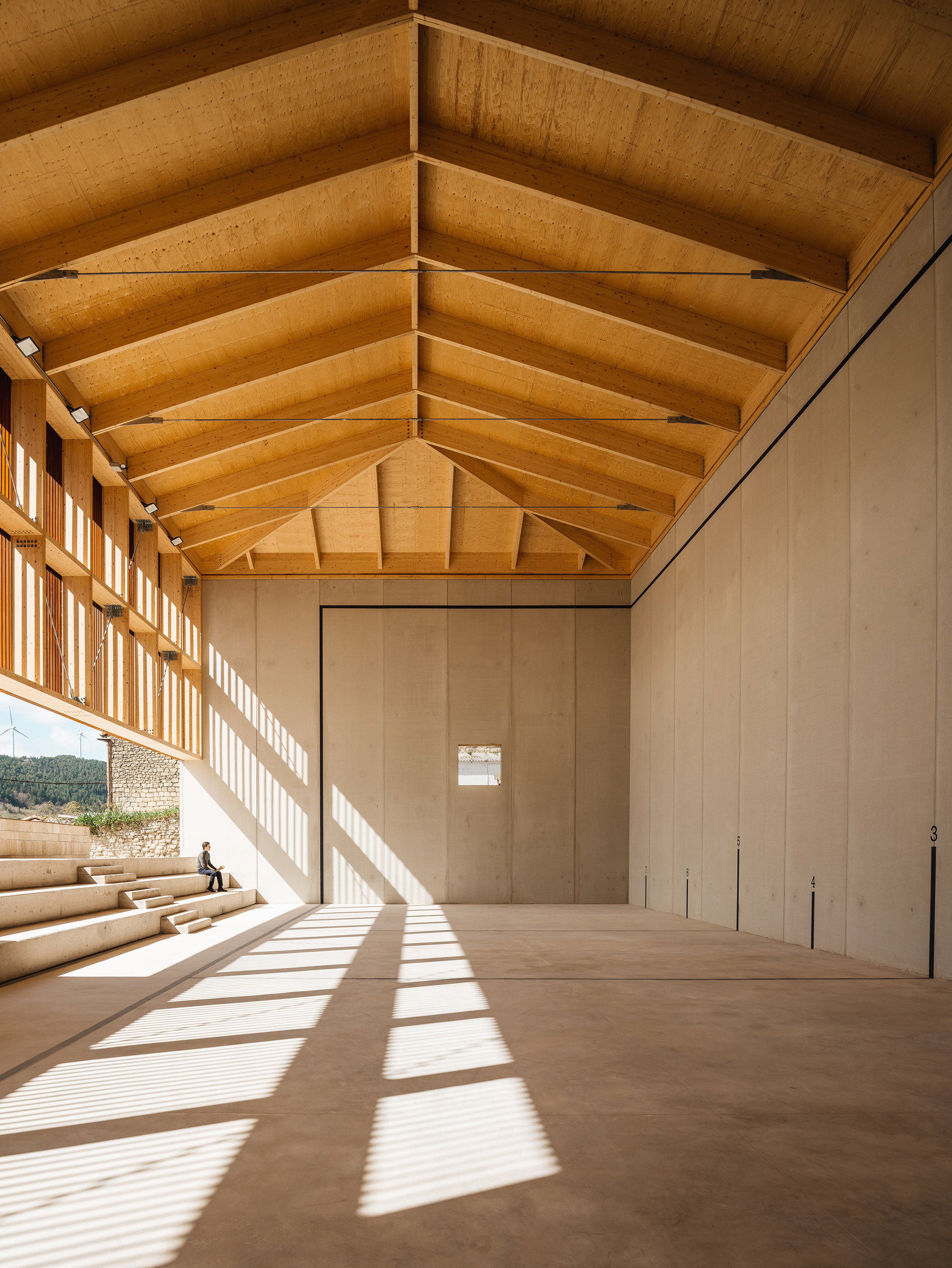 Striped sunlight from the slatted timber wall falls across the court floor, with a person seated on the concrete steps