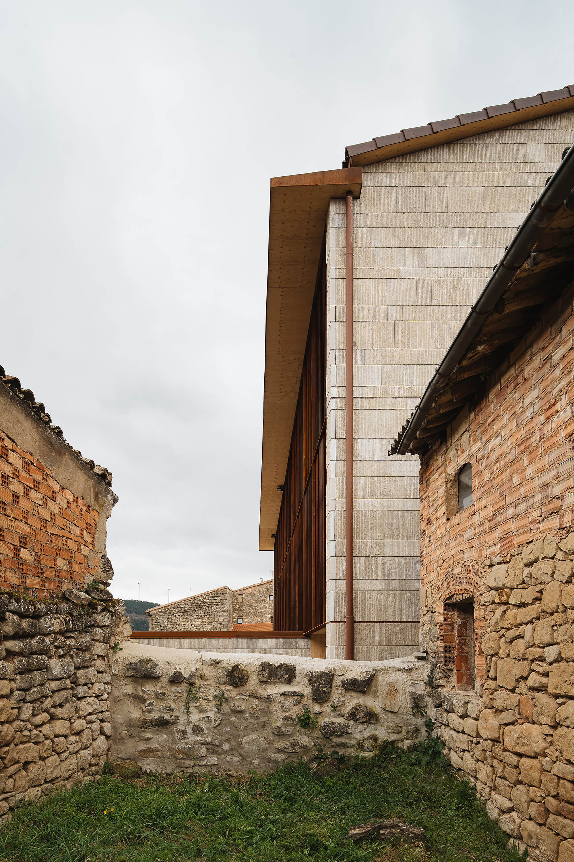 Gap between old brick and rubble stone walls framing a stone-clad building with copper downpipe under an overcast sky