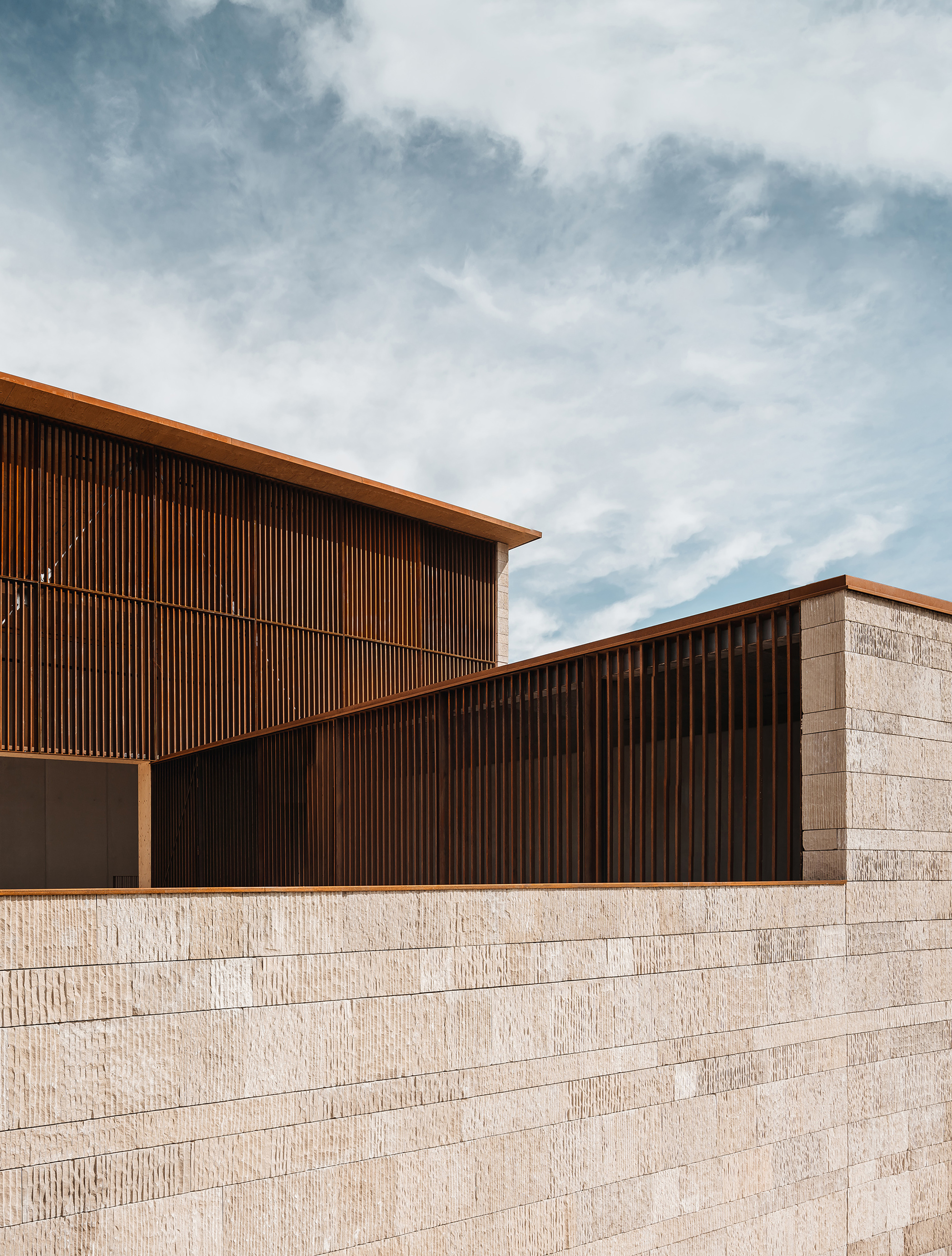 Rusted steel slat screens and thin cantilevered roof edges rising above a coursed limestone wall against a cloudy blue sky